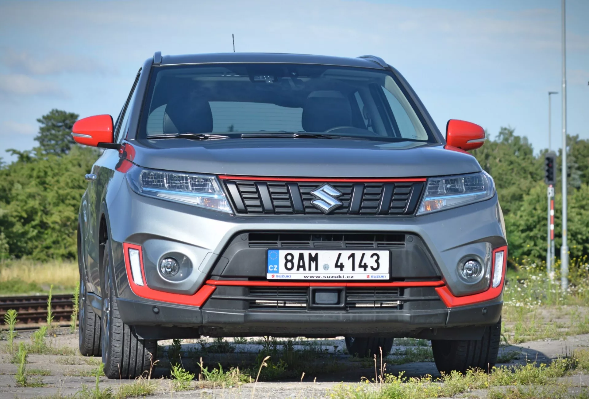 A képen egy modern 2018-as Suzuki Vitara SUV látható enyhén előre dőlő szögből, kiemelve az első részt és az utasoldal egy részét egy ritkás tájháttér előtt, amelyhez vasúti sínek tartoznak. Az autó látszólag kavicsos felületen parkol, és kiemelkedő, szögletes rácsot mutat be, elegáns fényszórókkal, valószínűleg LED vagy HID technológiával, valamint beépített nappali menetfénnyel vagy ködlámpákkal az első lökhárítóban. A lökhárítóban kontrasztos színű betét található, ami terepjáró vagy sportos kivitelre utal, és a látható hasmagasság megerősíti az SUV jellegét. A motorháztető emelkedett vonalai hozzájárulnak az agresszív stílushoz, míg az oldalsó tükrök és a sárvédők modern dizájnt mutatnak. A részben látható könnyűfém kerekek kiegészítik a robusztus megjelenést. A közeli felvétel lehetővé teszi, hogy részletesen megvizsgáljuk ezeket a jellemzőket.