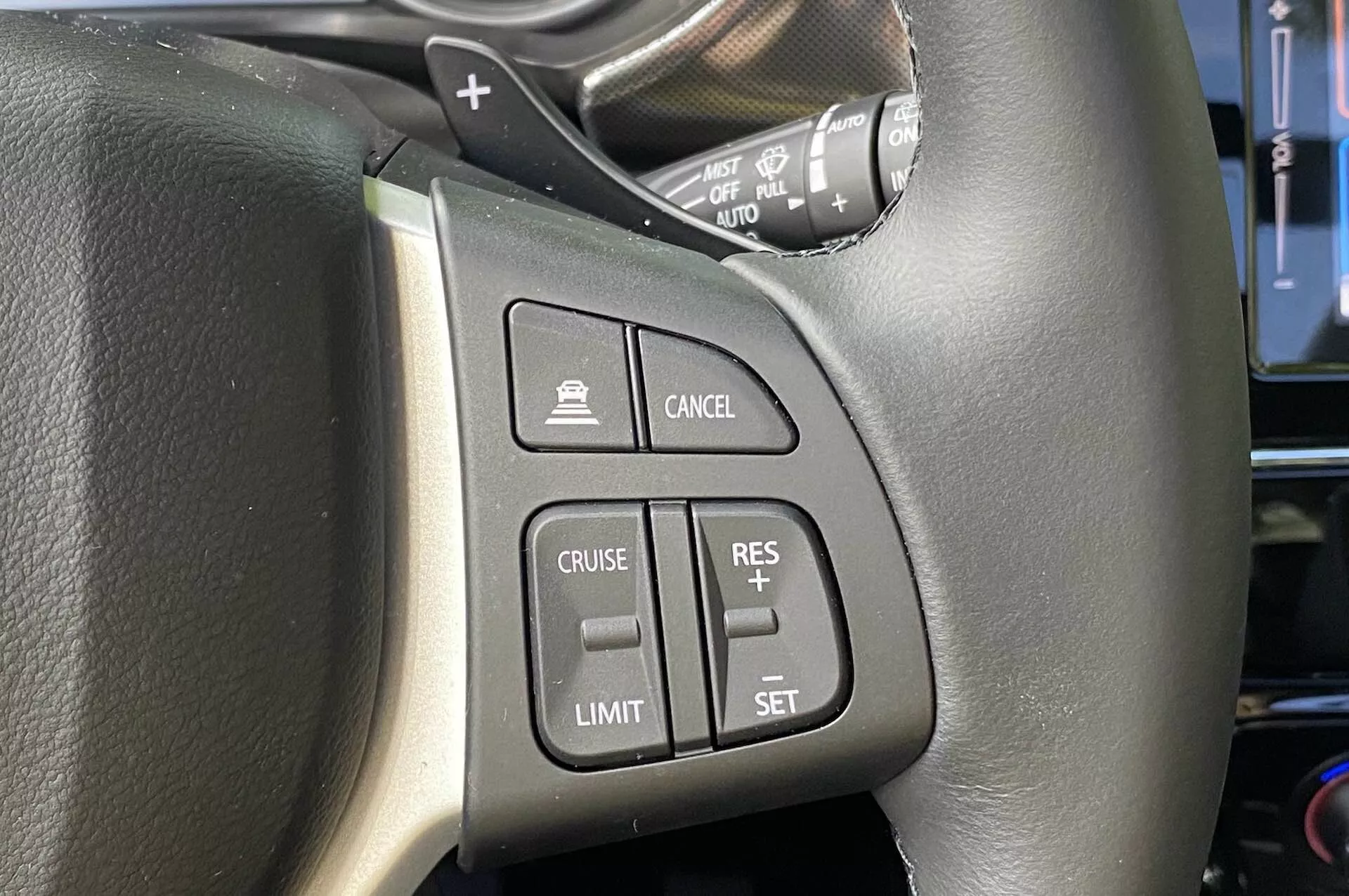 Közelkép a 2018-as Suzuki Vitara kormánykerékbe épített vezérlőiről, az elsősorban a bal oldalra összpontosítva. A fekete bőr kormánykerék látható varrással könnyen hozzáférhető gombokat tartalmaz, beleértve a sebességtartó automatika bekapcsoló gombját, a sebességkorlátozó bekapcsoló gombját, a sebességtartó automatika beállítási/kiigazítási gombjait és a törlés gombját. A képen egy vezérlőkar is látható, amely valószínűleg az irányjelzőkhöz és a fényszórókhoz tartozik, vezetői szemszögből nézve.