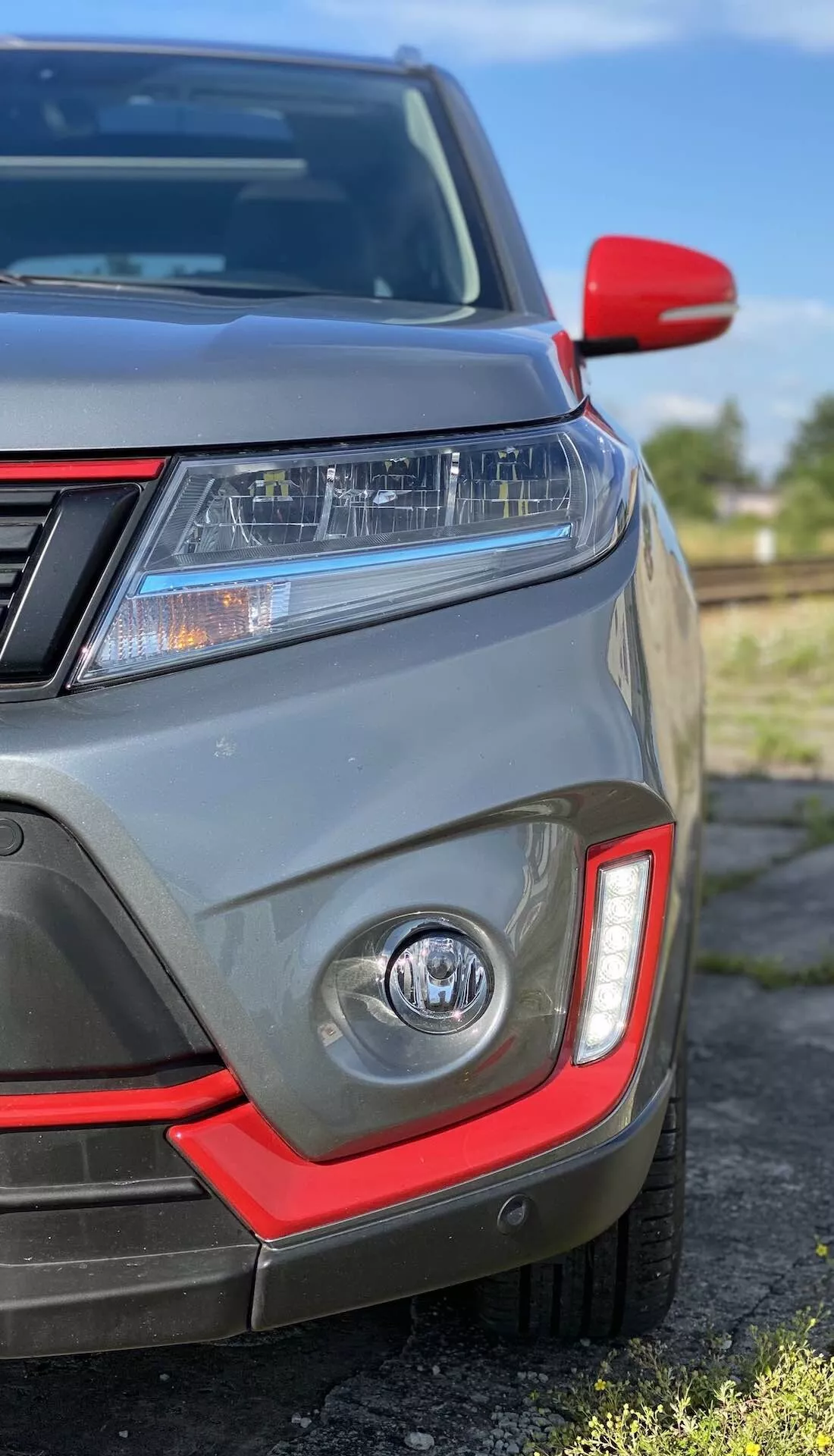 Közelkép a Suzuki Vitara 2018 bal első negyedéről, melyen LED fényszórók, nappali menetfények, piros akcentussal ellátott ködlámpa, parkolószenzorok, részleges első rács, piros kupakkal ellátott oldalsó tükör és metál szürke fényezés látható.