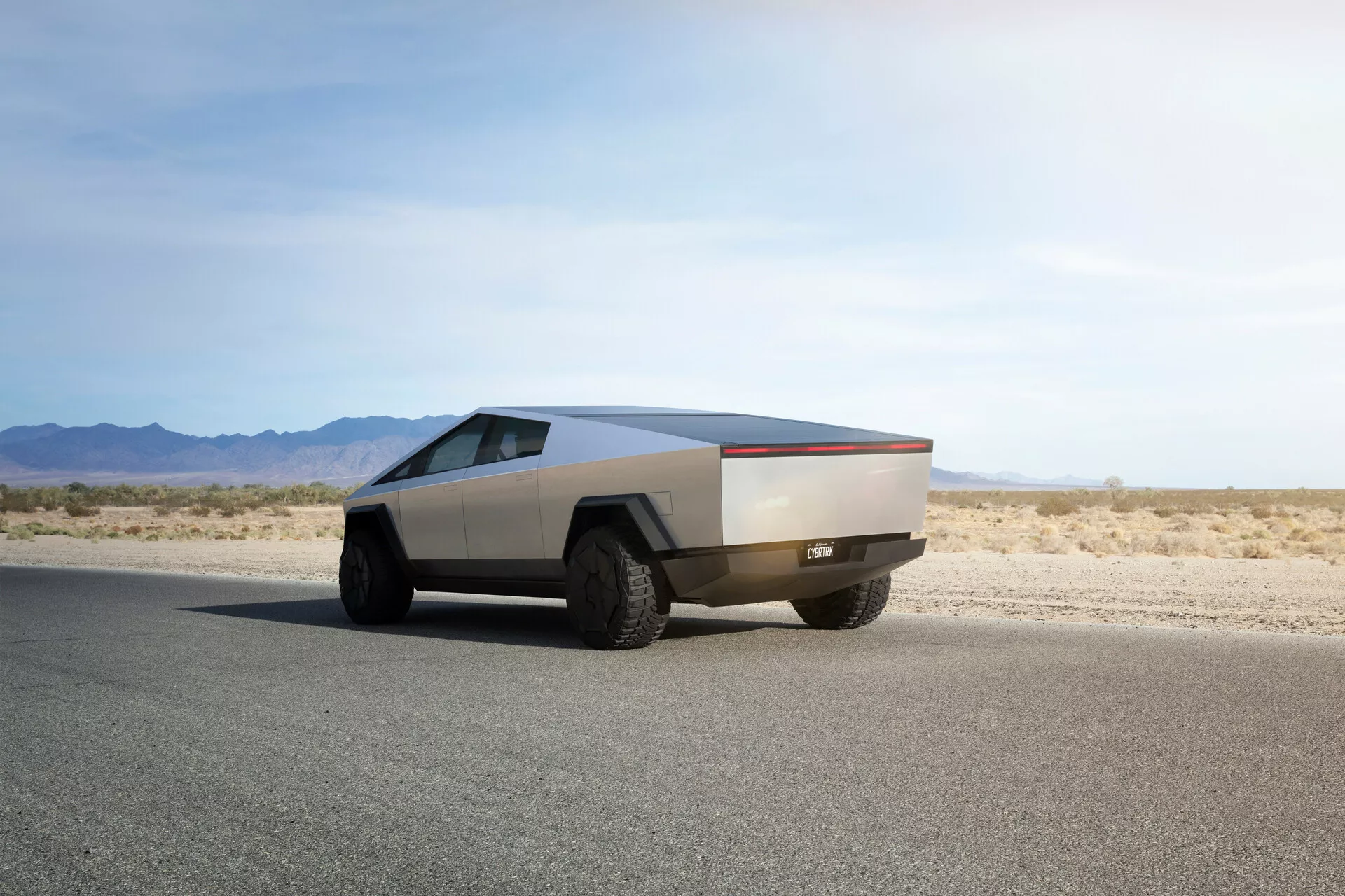 Futurisztikus Tesla Cybertruck 2021 sík aszfalt úton, hátsó háromnegyedes szögből rögzítve, mely kiemeli jellegzetes szögletes dizájnját, fémes felületét, nagy terepgumijait és a hagyományos tükrök és ajtókilincsek hiányát, ami fejlett automatizált mechanizmusokra utal.