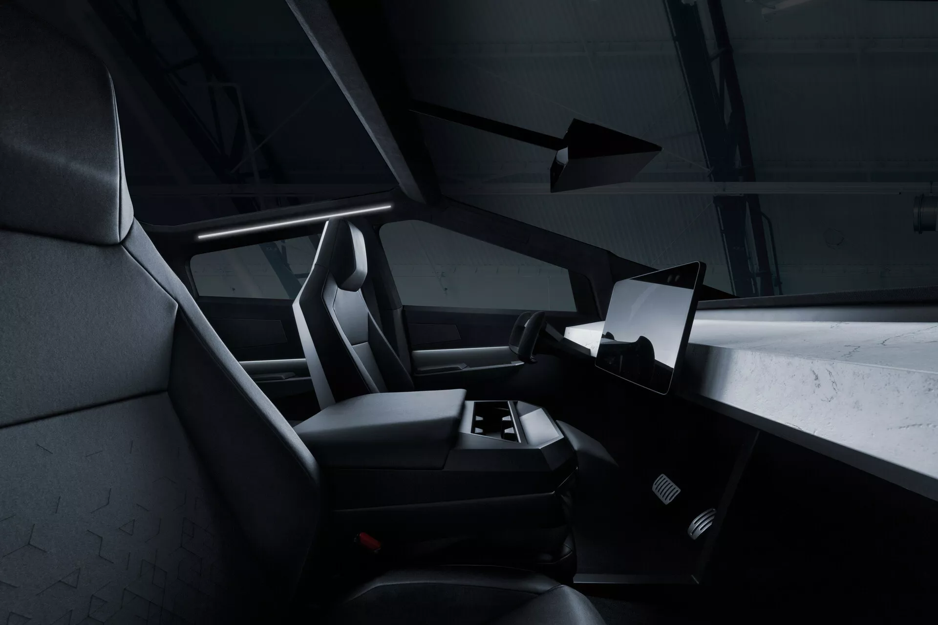 A Tesla Cybertruck 2021 (1. generáció) belső nézete a hátsó ülés nézőpontjából, bemutatva a kabin jobb oldalát, ahol egy nagy, vízszintesen orientált érintőképernyő dominál a műszerfal közepén. A kormánykerék egy része látható, de többnyire takarva van. Az első utas- és vezetőülések integrált fejtámlákkal és texturált kárpitozással rendelkeznek, valószínűleg szövetből vagy szintetikus bőrből. Az utastér lábrészén látható egy autószőnyeg, és nincsenek manuális vezérlők, ami minimalista, digitális megoldásokra utal. A sötét színvilág és a finom akcentusvilágítás modern, luxus hatást kölcsönöznek.