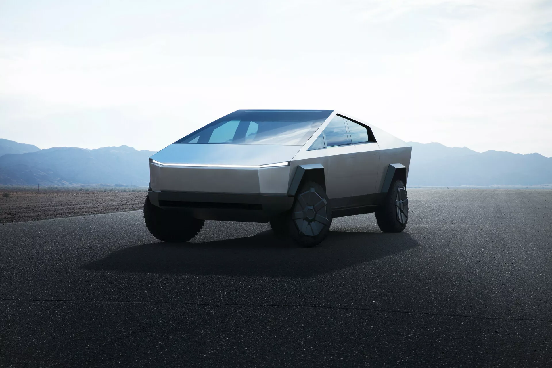 2021-es Tesla Cybertruck (első generáció) kép egy úton lapos tereppel vagy sivatagszerű hegyekkel a háttérben nappali fényben tiszta égbolttal. A fotó alacsony szögből készült, elölről és oldalról néző nézetet nyújtva, amely kiemeli a teherautó szögletes, futurisztikus dizájnját. A jármű ék alakú elejű, lapos panelekből készült karosszériával, vékony, vízszintes fényszórókkal, folyamatos lejtős szélvédővel, látható oldalsó tükrök nélkül, nagyméretű terepjáró kerekekkel bonyolult felnikkel és első rács nélkül, ami az elektromos hajtásra utal.