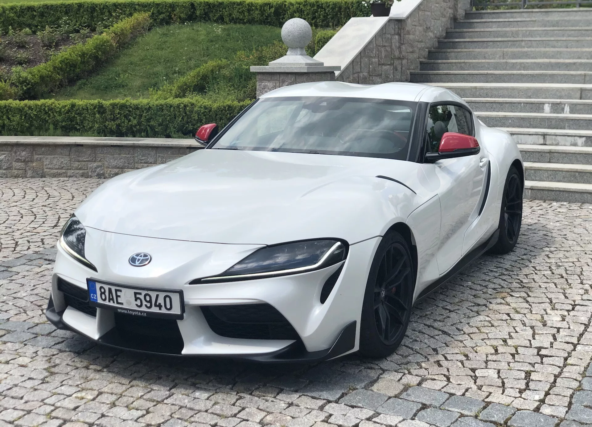 A képen egy 2019-es fehér Toyota Suprát (toyota-gr-supra-2-0-pure-automatic) láthatunk, háromnegyedes elölnézetből. Jól látható az autó alacsony és széles testtartása, éles szögletes fényszórói és fekete, többléces könnyűfém kerekei. A fotó kiemeli a karcsú dizájnt, az aerodinamikai kontúrokat és a teljesítményesztétikát. A sportautó járdán parkol, amelyen padkát és egy lépcsőt láthatunk a háttérben, ami városi vagy lakóövezeti környezetre utal.