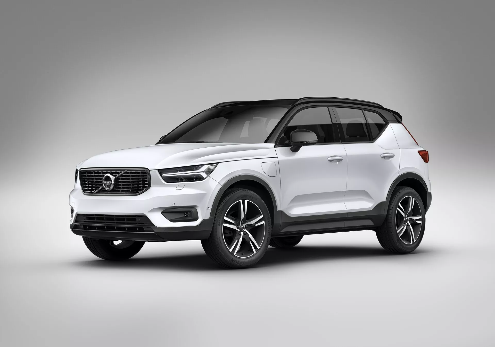Stúdiófotó egy fehér Volvo XC40 2018 (első generáció) modern SUV-ról. Alacsony szögből készült, a kép kiemeli az autó állását és dizájnelemeit, beleértve a háromnegyedes elülső nézetet, amely megmutatja az első és oldalsó profilokat. A fényszórók modern alakúak, esetleg LED technológiával, míg a hűtőrács tartalmazza a márka emblémáját és minimalista vízszintes vonalakat. Az alsó lökhárító ködfényszórókat vagy légbeömlőket integrál, jelezve a fejlett technológiát. Kiemelkednek a nagy, legalább 18 hüvelykes könnyűfém kerekek, a karosszériaszínű ajtókilincsek, tükörborítások és a panoráma napfénytető. A tetővonal hátulra keskenyedik, kupénszerűen, amely tovább fokozza a luxusérzetet, krómos vagy fémszegélyekkel az ablakok körül. A sima, fényes festékfelület és a stúdióhátter kiemeli a jármű tulajdonságait zavaró tényezők nélkül, biztonságra, kényelemre, infotainmentre és fejlett hajtásrendszerekre összpontosítva.