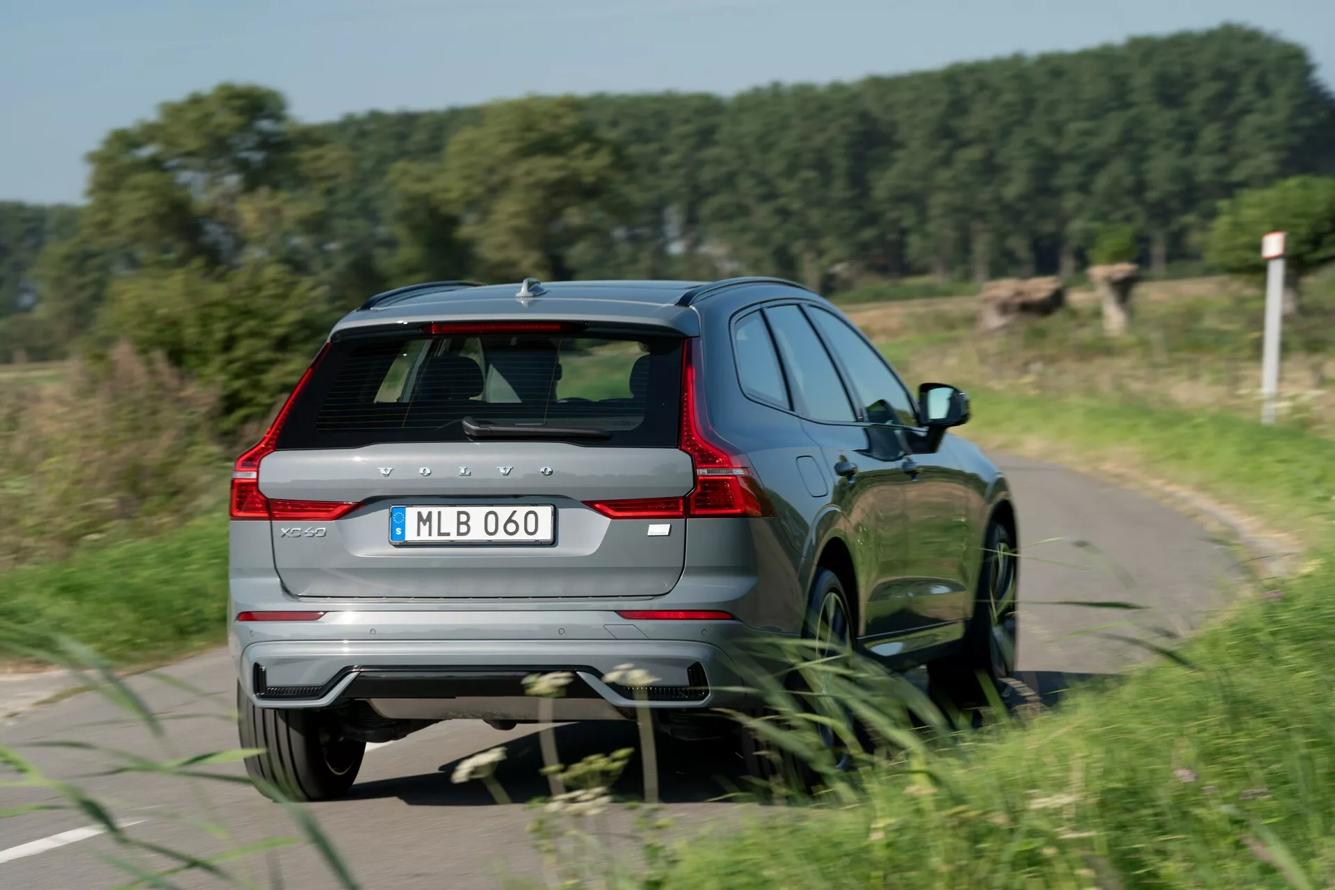 Háromnegyedes hátsó nézet egy 2021-es Volvo XC60-ról (második generációs faceliftről) mozgás közben, amely bemutatja a hátsó és jobb oldali dizájnelemeket, beleértve a hátsó ablaktörlőt, a spoilert és a harmadik féklámpát, a hátsó lökhárítót integrált fényvisszaverőkkel és kettős kipufogórendszerrel, meghosszabbított LED hátsó lámpákat, a jobb oldali tiszta profilját hátsó utasajtóval és többszörű küllős alufelnikkel, valamint a modell jelölését. Az autó egy keskeny, vidéki úton halad, a mozgás érzékelhetőségével, enyhe mozgáselmosódással a kerekeknél.