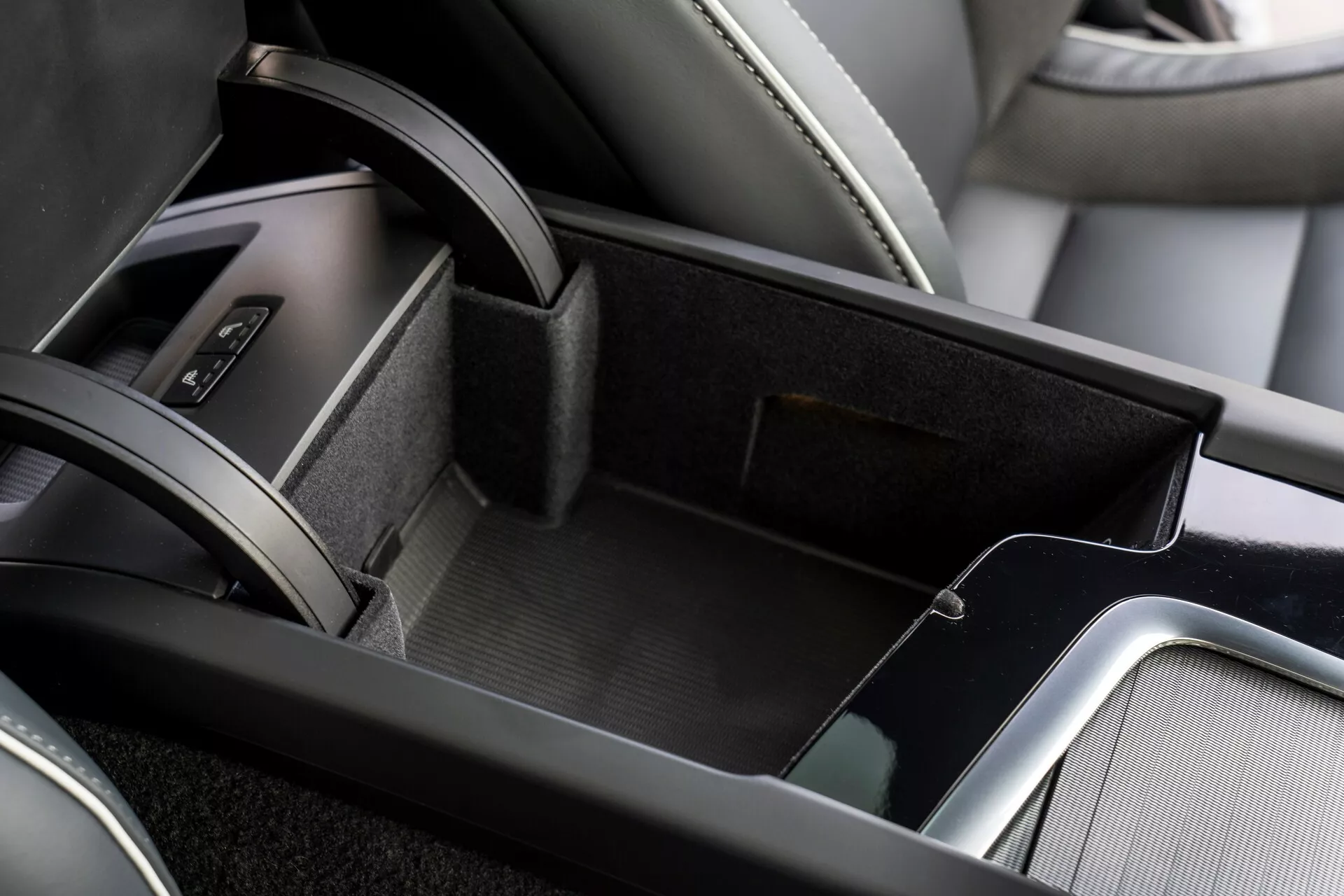 Közelkép a 2021-es Volvo XC60 belső terének középkonzol területéről, amely bemutatja a nyitott tárolórekeszt, amely filcszerű anyaggal van bélelve, két szinten elhelyezett tárolót kisebb felső tálcával, bőrrel bevont könyöktámaszt és fémes díszítéseket a kifinomultság növelése érdekében. A kép szélén látható a sebességváltó terület egy része bőr gumiharanggal.