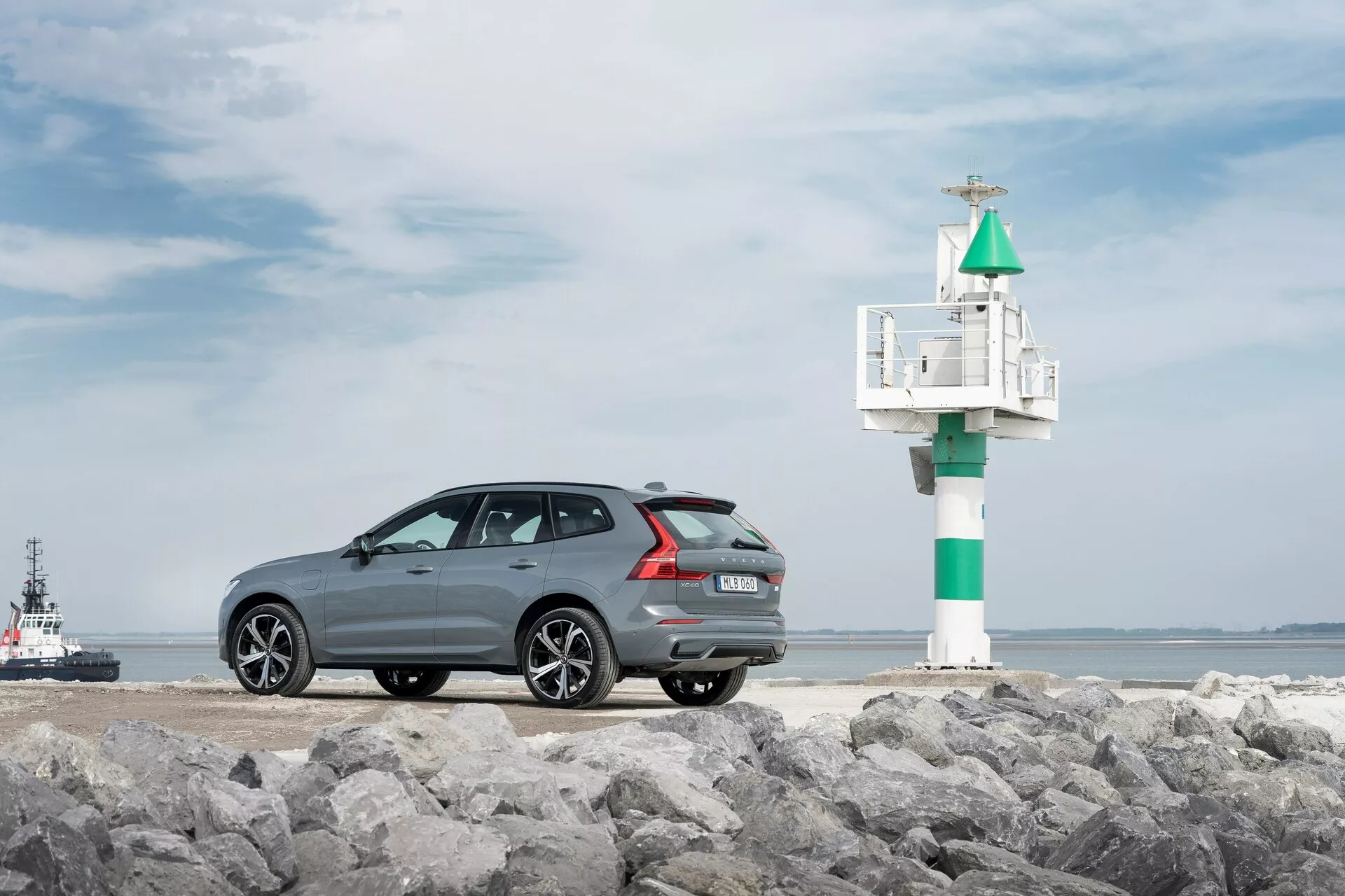 A Volvo XC60 2021 második generációs, felfrissített modelljének fényképe, amely háromnegyedes hátsó szögből készült egy sziklás felületen, tengerparti háttérrel. A sötétszürke, metálfényű autó sportos, elegáns tetővonallal, többrudas könnyűfém felnikkel, hátsó parkolószenzorokkal és dupla kipufogóval rendelkezik. Az autó jobb oldalán egy kis világítótorony vagy jelző van látható, a vízben egy vontatóhajóval vagy pilótahajóval, amely fokozza a part menti hangulatot.