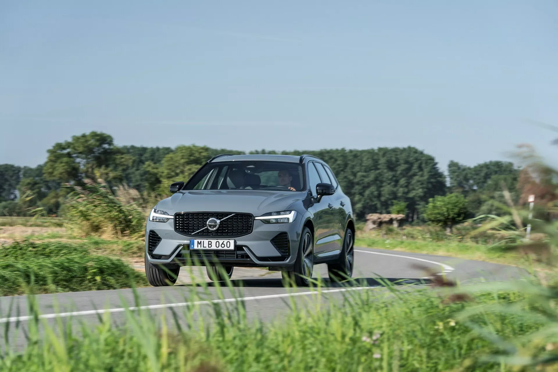 Modern SUV, Volvo XC60 2021 (2. generáció facelifttel), alacsony elülső háromnegyedes szögből elkapva, kiemelve a dinamikus tartást és a dizájn jellemzőit. Jellegzetes rácsos kialakítás nagy emblémával, elegáns LED fényszórók és formázott lökhárító beépített légbevezető nyílásokkal. Aerodinamikus, kifinomult megjelenés kontúrozott vonalakkal, magas hasmagasság, nagy többrudú könnyűfém kerekek, alacsony profilú abroncsok, integrált irányjelzővel ellátott oldalsó tükrök, lejtős tetővonal és sötétített ablakok. Modern technológiai és biztonsági funkciók jelenlétére utal, vidéki úton közlekedve, elmosódott növényzettel, amely a mozgást jelzi.