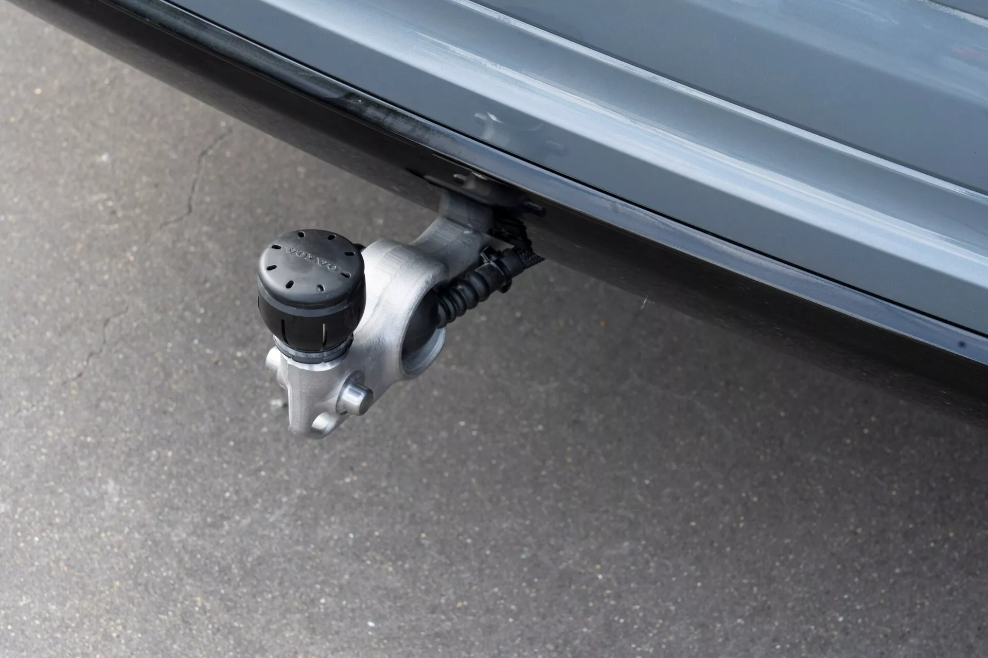 Közelkép egy 2021-es Volvo XC60 (2. generáció-felújított változat) kihúzható oldalfokáról, amely az ajtó alatti területen egy fémből készült zsanérmechanizmushoz van rögzítve. Az alacsony szögből készült fotón jól látható a biztonságos belépést segítő, csúszásmentes, kiemelt fekete pontokkal ellátott felület, amely automatikusan kinyílik és becsukódik.