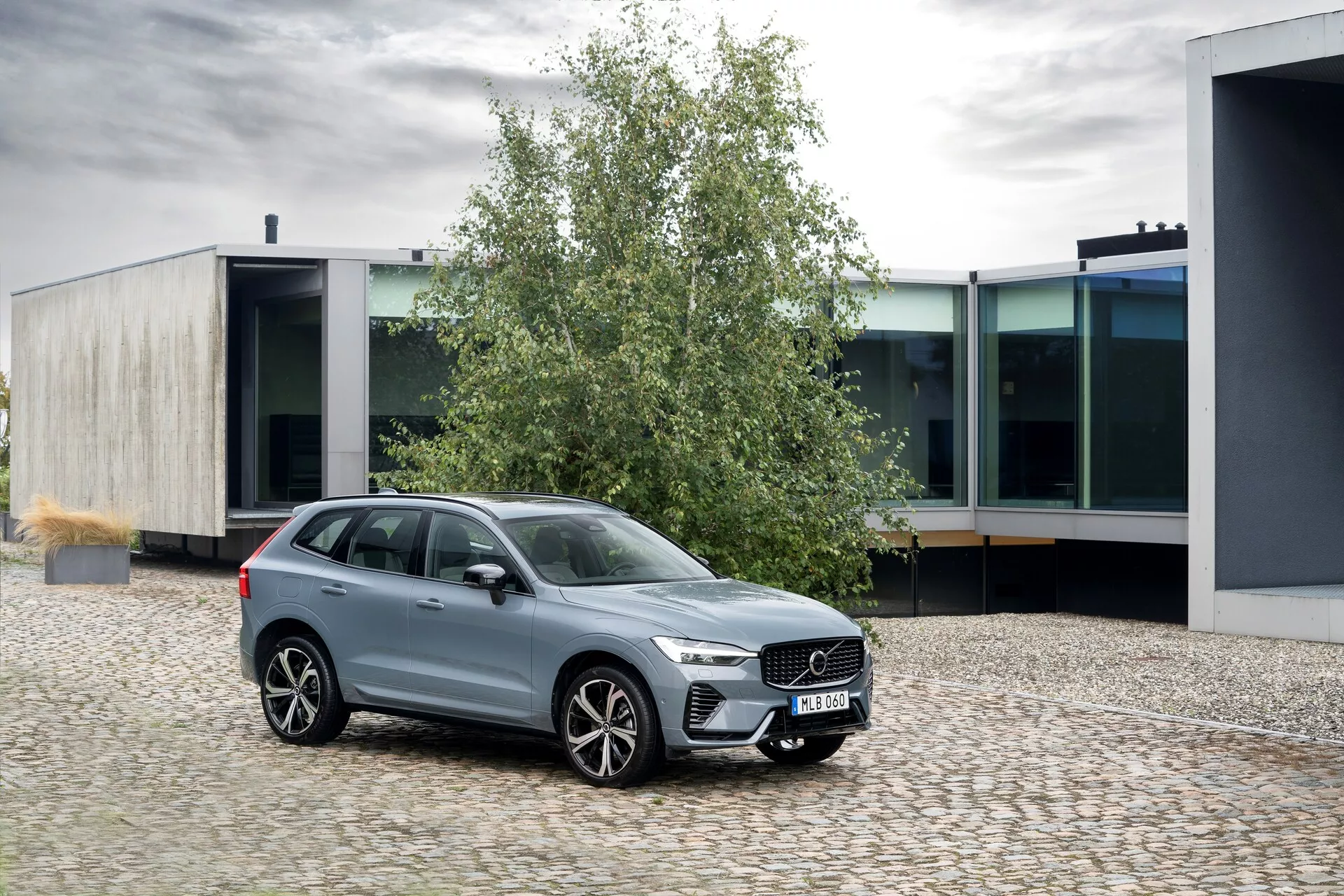 A képen egy 2021-es Volvo XC60 (2. generáció-facelift) látható egy macskaköves felületen parkolva, enyhe szögben balra néző helyzetben. Az elülső háromnegyedes nézet kiemeli a prominens függőleges rácsot, a karcsú LED fényszórókat nappali menetfénnyel, valamint a nagy, többrészes alufelniket. Az ablakokat króm borítás veszi körül, ami fokozza a polírozott fémes megjelenést. Az SUV megemelt hasmagassággal, tetősínekkel és karosszéria színű visszapillantó tükrökkel rendelkezik beépített jelzőfényekkel. A jelenet modern építészettel rendelkezik egy városi környezetben, kiemelve az autó elegáns dizájnját és luxus jellemzőit.