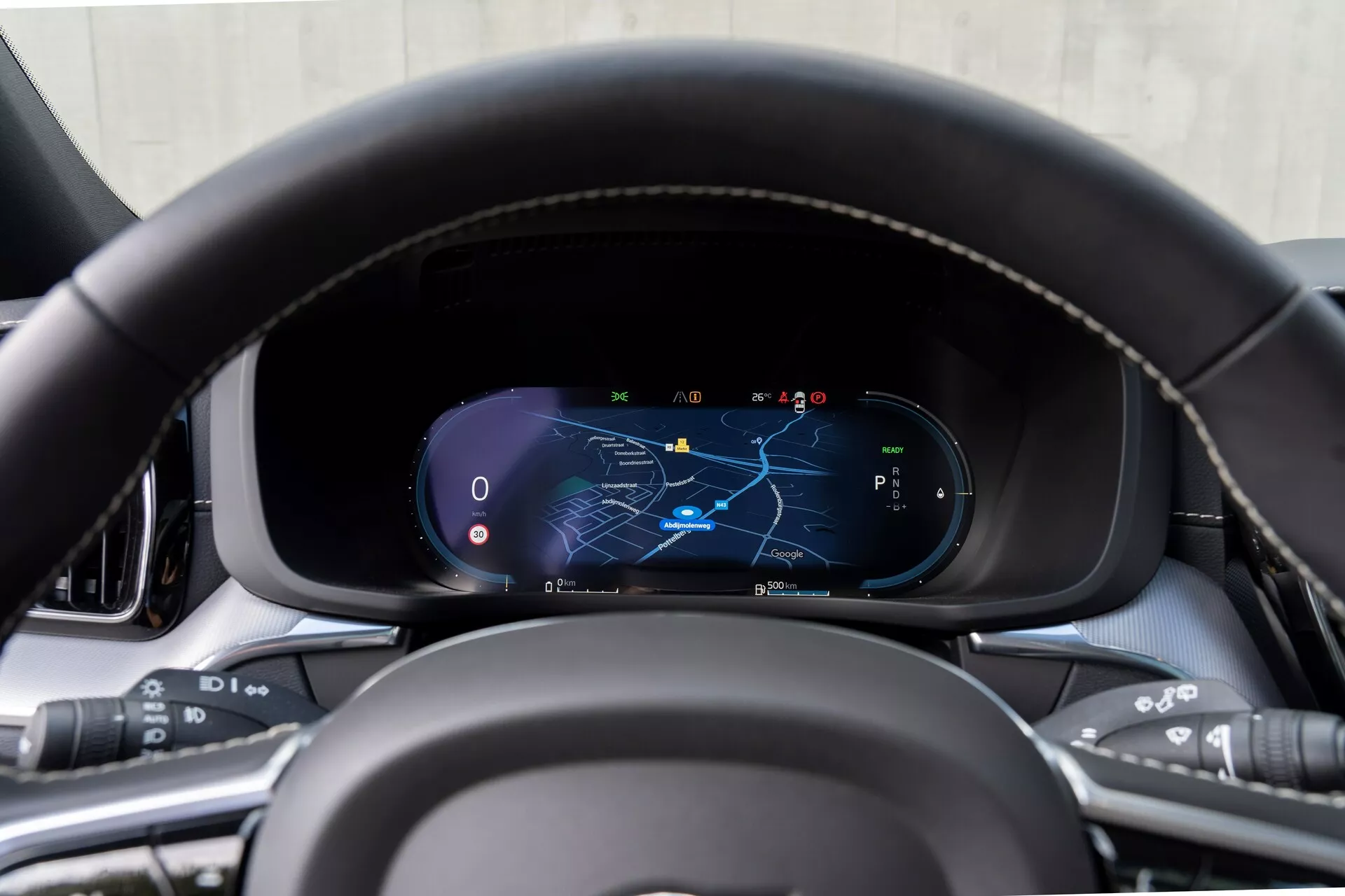 Közeli kép a 2021-es Volvo XC60 (2. generáció-felújítás) műszerfaláról a vezetőülés perspektívájából, fókuszálva a digitális műszercsoportra, bőrhatású, varrott kormánykerékkel, amelyen többfunkciós gombok találhatók a médiakezeléshez, hangutasításokhoz, tempomathoz és vezetői segédrendszerekhez. A digitális kijelző térképet mutat kiemelt navigációs útvonallal, sebességmérővel, fordulatszámmérővel és parkoló üzemmódban lévő sebességváltó kijelzéssel, prémium belső anyagokkal körülvéve.