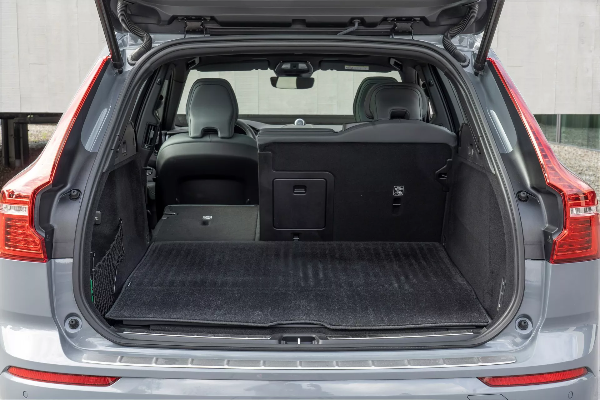 A kép a 2021-es Volvo XC60 (2. generációs arcplasztika) hátsó csomagterét mutatja, szemből nézve, nyitott csomagtérrel. A középtávú felvétel a csomagtér teljes szélességét lefedi, sík rakodófelülettel, amely fekete szőnyegszerű anyaggal van bevonva. A bal oldalon egy kis tárolórekesz hálóval található. A jobb hátsó ülés lehajtott, felfedve az osztott összehajthatóságot, míg a bal ülés függőleges helyzetben marad, fejtámlával. A hátsó ülések felett egy visszahúzható csomagtértakaró található, amely a jármű tárolási sokoldalúságát szemlélteti.