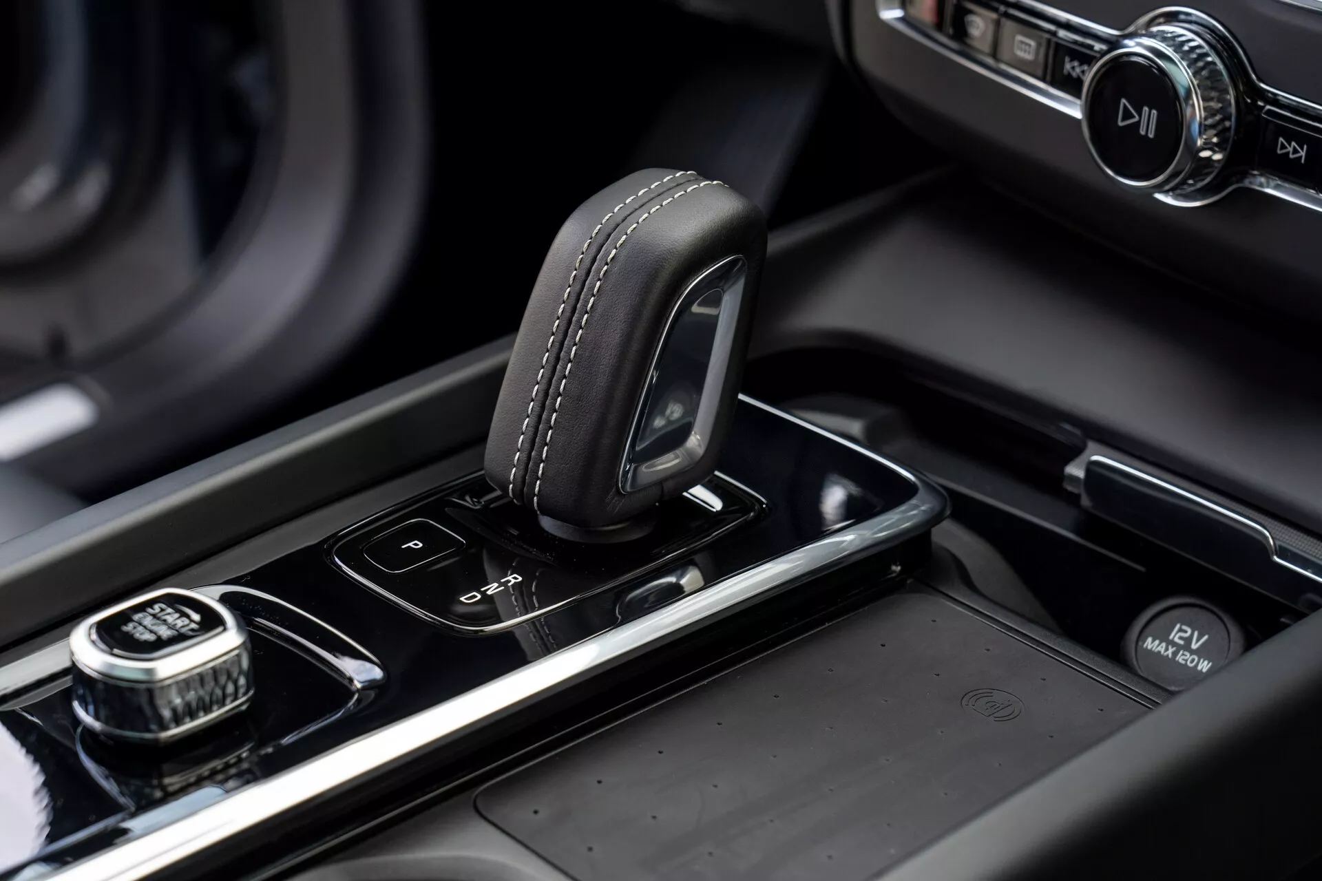 Közelkép a 2021-es Volvo XC60 (második generációs, faceliftes) középkonzoljáról. A kép kihangsúlyozza a bőrből készült, varrási részletekkel rendelkező, elektronikus váltókart egy fényes fekete felületen. Jobb oldalon egy recézett szélű tárcsa látható, valószínűleg az infotainment rendszer navigálására, nyomógombos kiválasztási funkcióval. A váltókar mellett parkolószenzorok és automatikus start/stop rendszer gombjai találhatók. A középkonzol fém díszítéseket is tartalmaz, és a háttérben egy megvilágított infotainment képernyő látható, hangsúlyozva a jármű modern, prémium színvonalú kialakítását és kivitelezését.