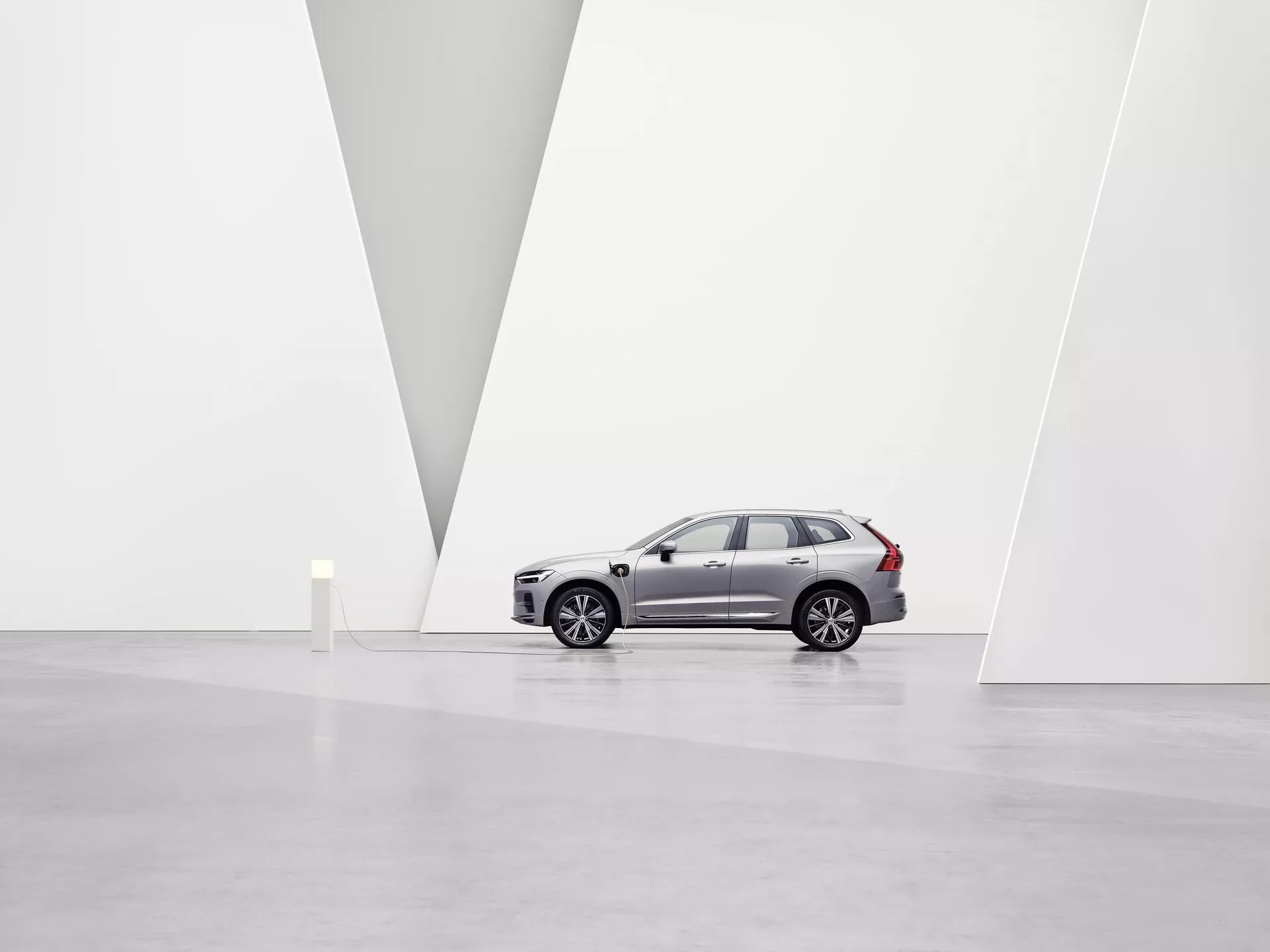 A képen egy Volvo XC60 2021 második generációs ráncfelvarrott SUV látható, tágas, minimalista művészeti galériában, fehér falakkal és fényes padlóval. A jármű balra néző, profilból látható, kiemelve az alufelniket, az ajtók karaktervonalait, az oldalsó tükröket, a hátsó ablakok sötétített üvegét és a jellegzetes tetősíneket. Az enyhén alacsony szögből látható az első hűtőrács, a LED fényszórók, az integrált irányjelzők és az első lökhárító parkolószenzorai. A tiszta környezet és a fal V alakú válaszfala kiemeli az autó dizájnját és prémium piaci pozícióját a lágy, szórt fényviszonyok alatt.