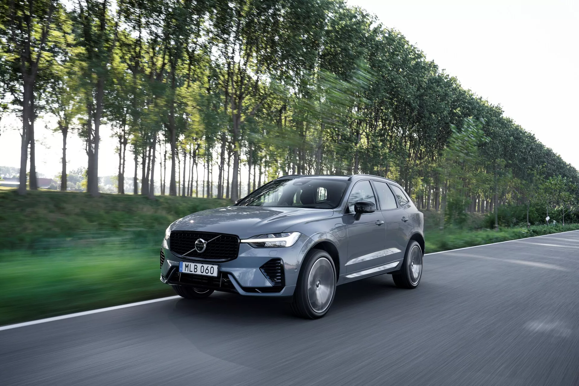 A képen egy mozgásban lévő Volvo XC60 2021 (2. generációs faceliftes) látható, háromnegyedes elülső szögből megörökítve, amelyen jól kivehető a jármű vezető oldala. Ez a modern luxus SUV elegáns kialakításával és az első hűtőrácson látható emblémával tűnik ki. Az enyhén lefelé irányuló szögben készített felvétel magában foglalja a motorháztetőt, a lökhárítót, a hűtőrácsot, a fényszórókat és a kerekeket. A sötét metál színű, feltehetően szürke vagy fekete jármű fényes kivitelben, könnyűfém kerekekkel, LED fényszórókkal és aerodinamikus karosszériával rendelkezik. Az elmosódott háttér, amelyen fákkal szegélyezett utcarészlet látható, a mozgást sugallja, kiemelve az autó teljesítményét.