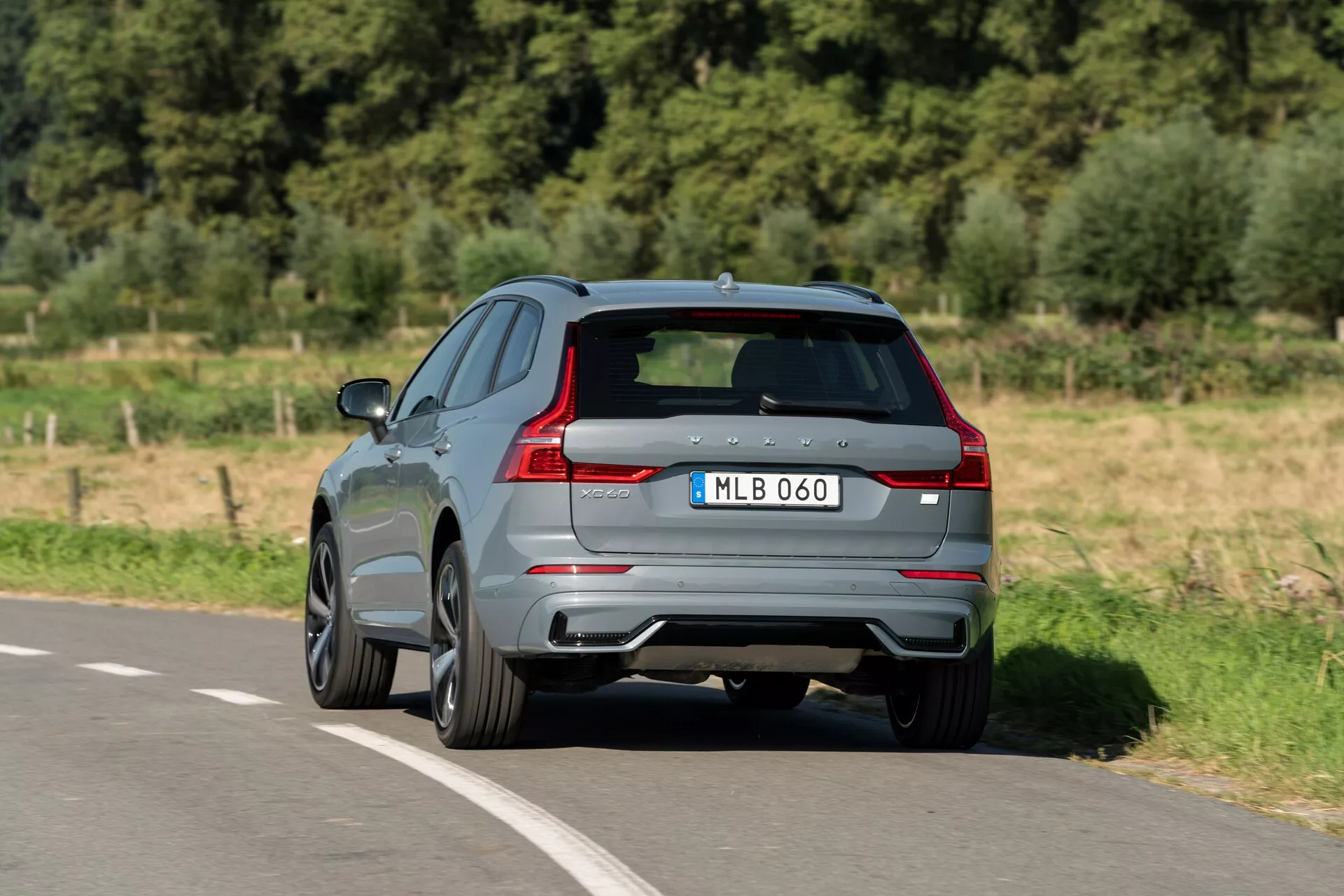 Háromnegyed hátsó nézet egy 2021-es Volvo XC60-ról, amely aszfaltozott úton halad. Egy emelt szögből rögzített, modern SUV látható fényes szürke festékkel, jellegzetes LED hátsó lámpákkal, dupla kipufogóvéggel és hátsó diffúzorral. A gyártó emblémája és a modell megnevezése látható a formázott hátoldali ajtón, sötétített ablakokkal és többküllős felnik dizájnnal, ami sportosabb változatra utal. A kép tiszta betekintést nyújt az aerodinamikai dizájnba és a modern járművekre jellemző prémium jellemzőkbe.