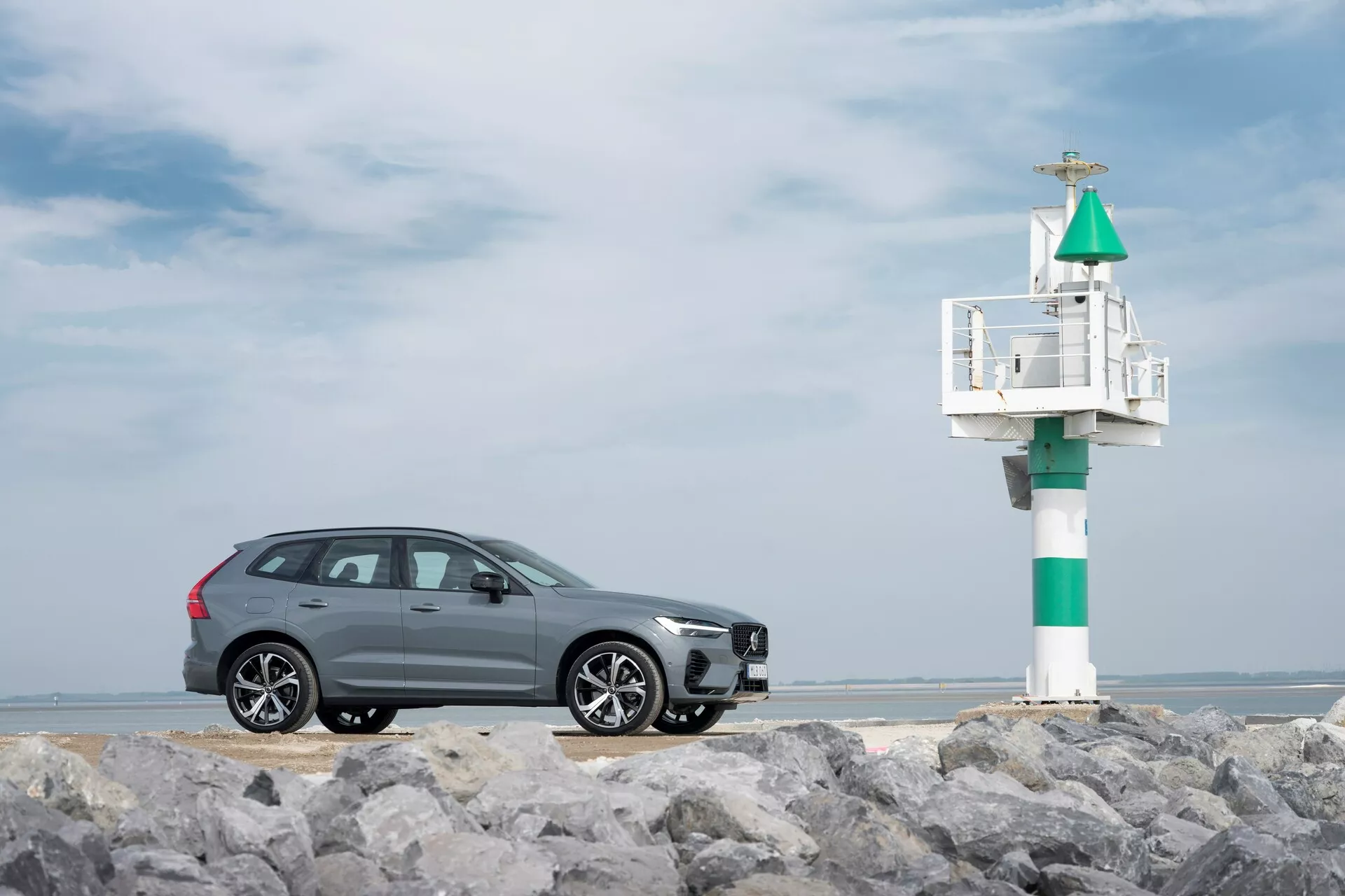Profil nézet egy szürke 2021-es Volvo XC60 (második generáció, facelift) SUV-ról köves talajon, tengeri világítótoronnyal a háttérben. Az elegáns dizájn króm ablakkereteket, többküllős alumínium felniket és integrált irányjelzőt tartalmaz az oldalsó tükörben, részben felhős tengerparti égbolttal a háttérben.