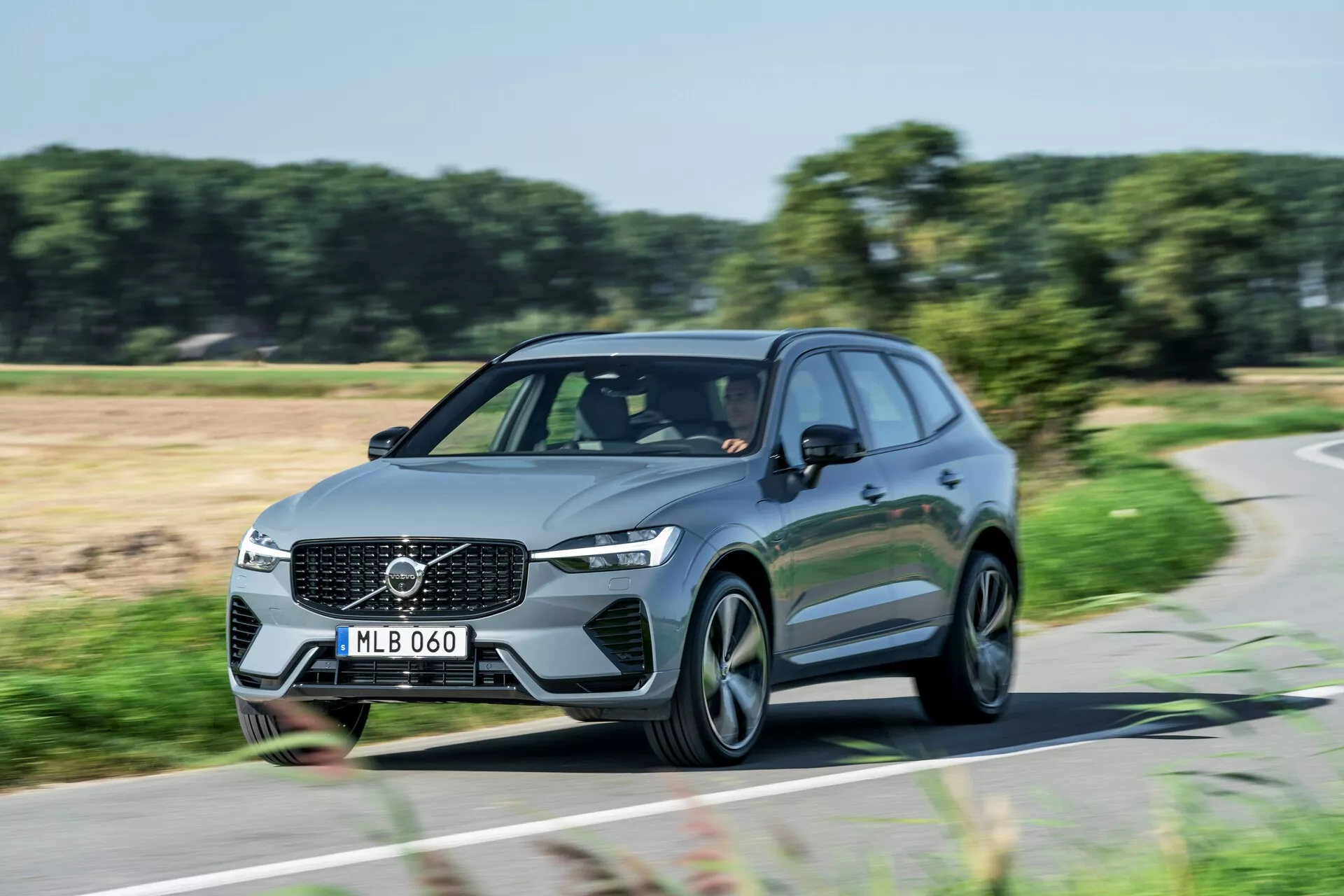 Dinamikus nappali felvétel egy mozgásban lévő Volvo XC60 2021 (2. generáció-facelift) SUV-ról, háromnegyed elölnézetből. A képen látható az első hűtőrács a Volvo emblémával, szögletes LED fényszórók, aerodinamikus vonalak és modern, többrudas alufelnik. A nappali menetfények világítanak, jelezve, hogy az autó mozgásban van. A fotó kiemeli a modern családi vagy luxusautó elegáns dizájnját és jelenlétét az úton.