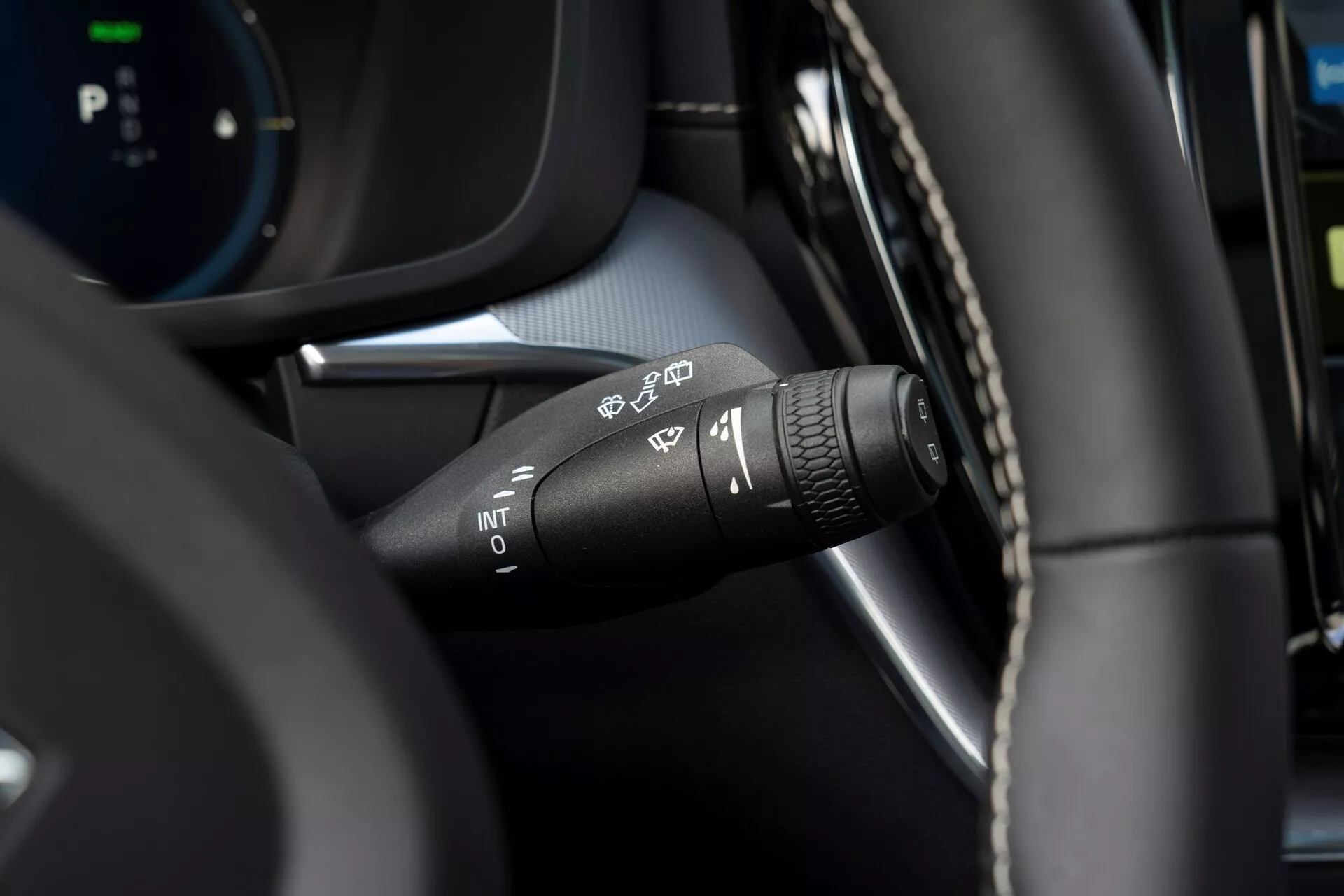 Közelkép egy 2021-es Volvo XC60 (2. generációs facelift) szélvédő-törlő kapcsolójáról a vezető szemszögéből. A különböző törlőbeállítási szimbólumokra összpontosítva: automatikus esőérzékelés, szakaszos, alacsony és magas sebesség. A kapcsoló végén egy forgatható gyűrű található a szakaszos törlési intervallumok beállításához. Modern belső tér, amely bőr jellegű anyag és fekete műanyag alkatrészek kombinációját tartalmazza, elhomályosult digitális műszerfallal a háttérben.