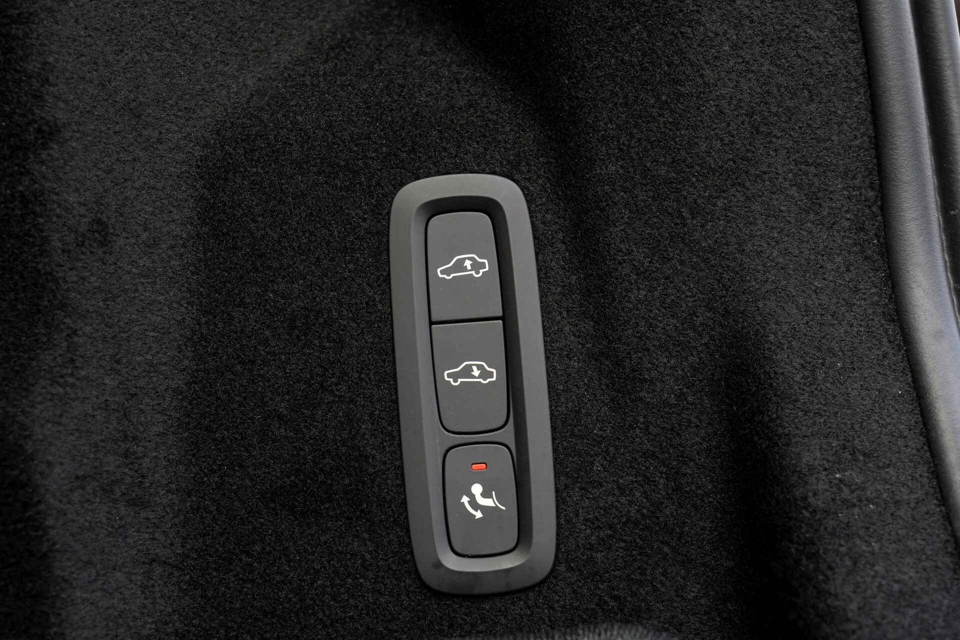 Közelkép egy vezérlőpanelről egy Volvo XC60 2021-es (2. generáció, faceliftes) modellben, amely műanyag burkolatba van beépítve fekete, szőnyegszerű háttér előtt. Négy gomb van rajta: felül - jármű nyitott csomagtartó-fedéllel, második - autó sziluettje nyitott ajtókkal, harmadik - autó egy nyíl mutat a motorháztető felé, alul - autó egy ívelt vonallal fölötte, amely valószínűleg csomagtartó nyitására, ajtózár/kireteszelésre, motorháztető-hozzáférés/valet mód és biztonsági rendszer aktiválására vagy pánikriasztásra utal.