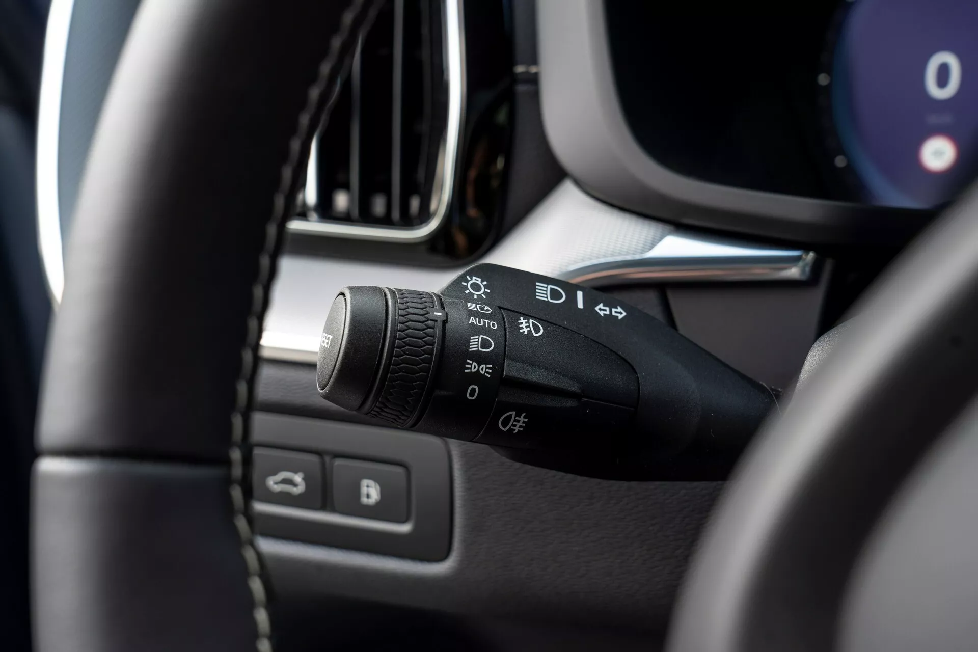 Közelkép a Volvo XC60 2021 (második generáció, ráncfelvarrott) belsejéről, a bal oldali kormányoszlopra összpontosítva. A fő téma a fényszóró és az irányjelző vezérlőkarja, különböző szimbólumokkal az automatikus világítás, parkolófények, fényszórók, ködlámpák beállításai és egy testreszabható funkció számára. A kép a vezetőülésből készült, kissé felfelé és balra nézve, az elmosódott kormánykerék bal felső részét mutatva az előtérben, hangsúlyozva a vezérlőkart. További elmosódott részletek közé tartoznak a műszerfal, a középkonzol, a légbeömlő vagy az infotainment képernyő kerete és a vezető oldali ajtó ablak- és tükörvezérlői.