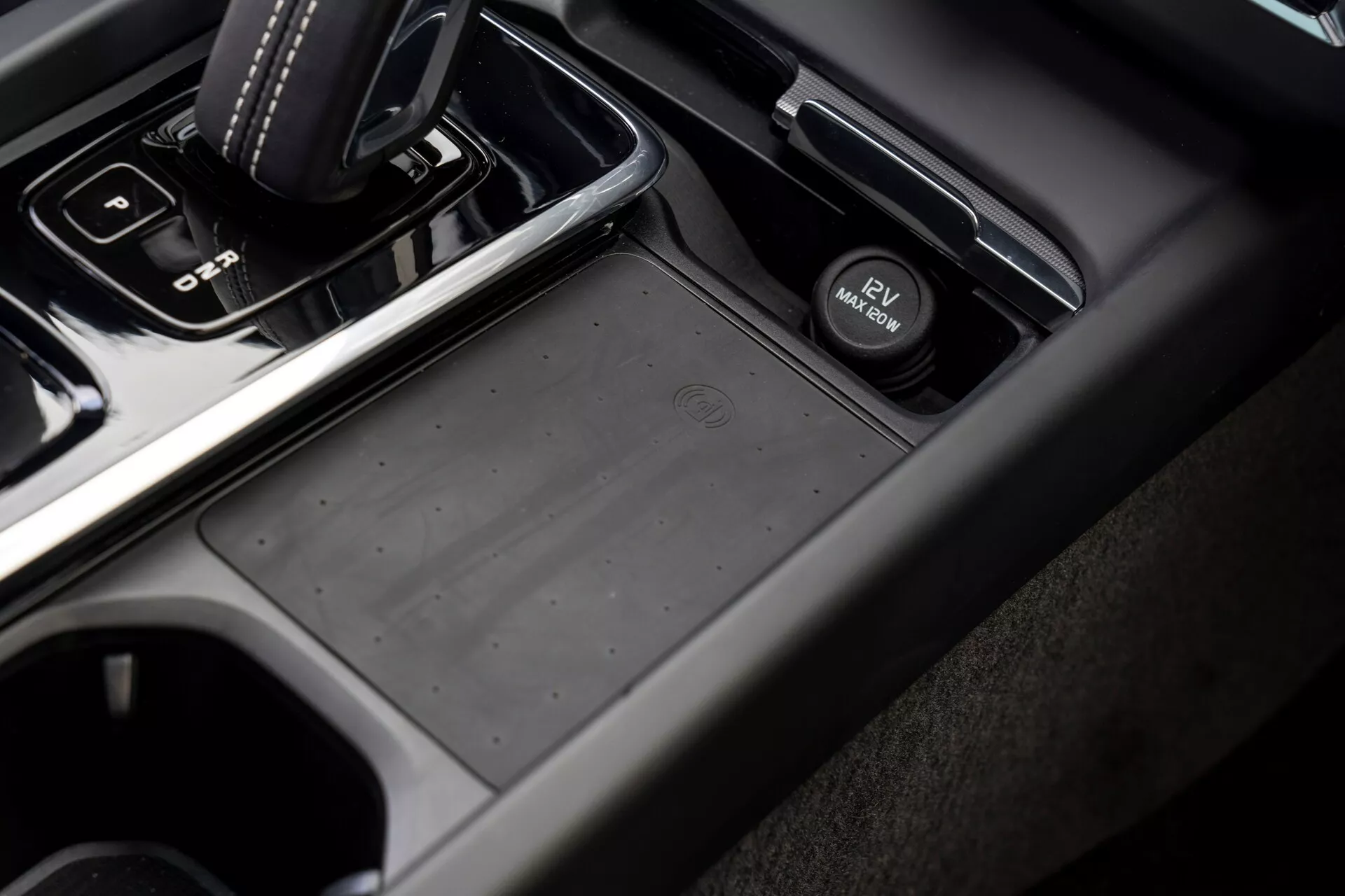 Közelről és szögben mutatja a Volvo XC60 2021 (második generáció, frissítés) középkonzolját az utas oldaláról, automata váltókarral 'park' pozícióban, csúszásmentes tárolórekesszel, elektronikus rögzítőfékkapcsolóval, pohártartókkal és sötétszürke és fekete tónusú, puha tapintású műanyag és fémes felületű anyagok kombinációjával.