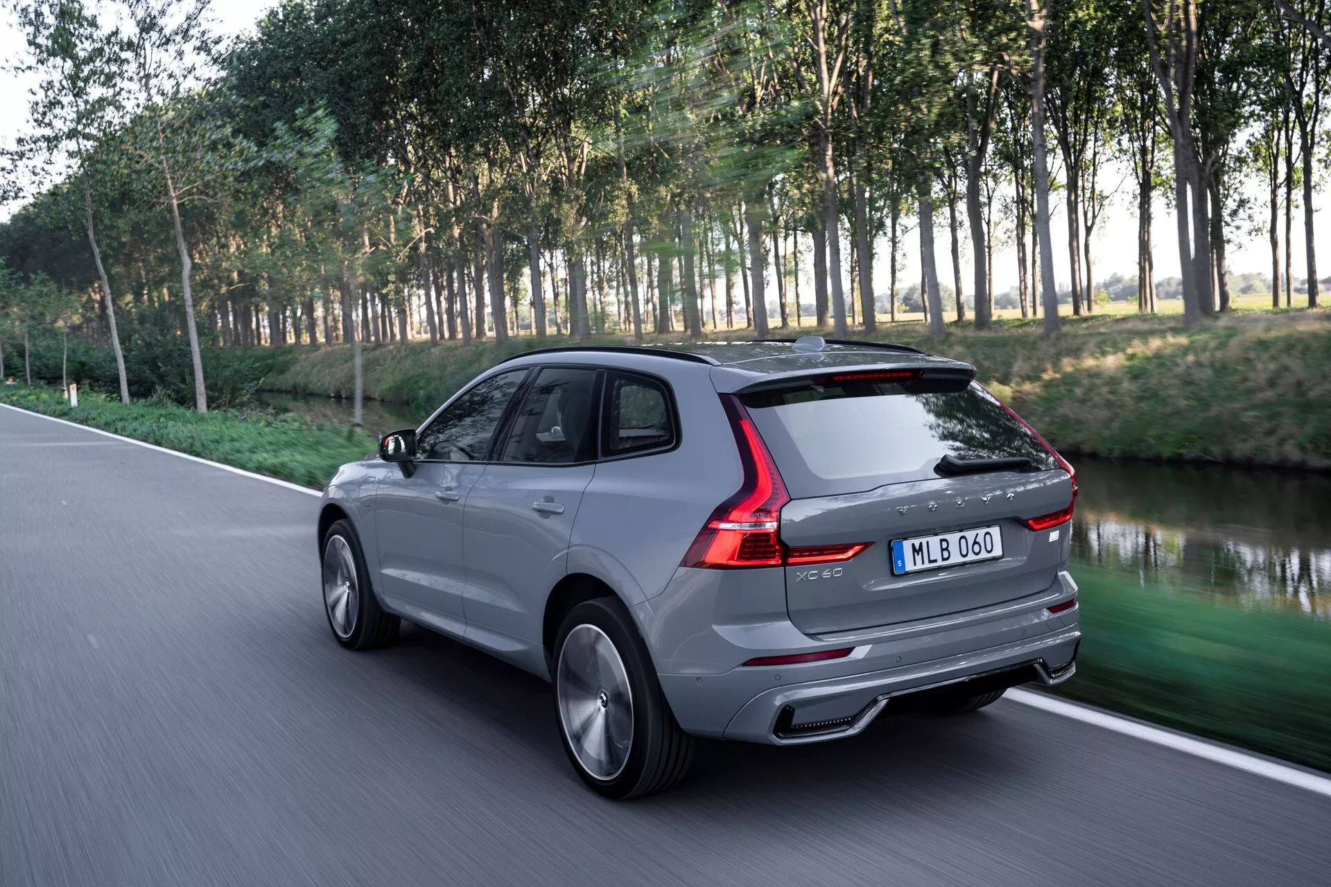 Dinamikus hátsó háromnegyedes nézet egy modern Volvo XC60 2021 (2. generáció-felújított) SUV-ról aszfaltúton, kiemelve az elegáns hátsó dizájnt, beleértve a hátsó lámpákat, a hátsó ablakot, a csomagtartót és a tetővonalat. Az alulnézetből, de az SUV középvonalánál alacsonyabb perspektívából készült kép stílusos kerekeket és oldalvonalakat mutat be. A SUV mozgásban van, elmosódott természetes háttérrel, fák és fű, valószínűleg park út mellett. Az ezüst-szürke színű luxus jármű kettős kipufogóval, aerodinamikus dizájnnal és fényes kivitelben van, alufelnik és valószínűleg fejlett technológiával, mint például LED hátsó lámpák.