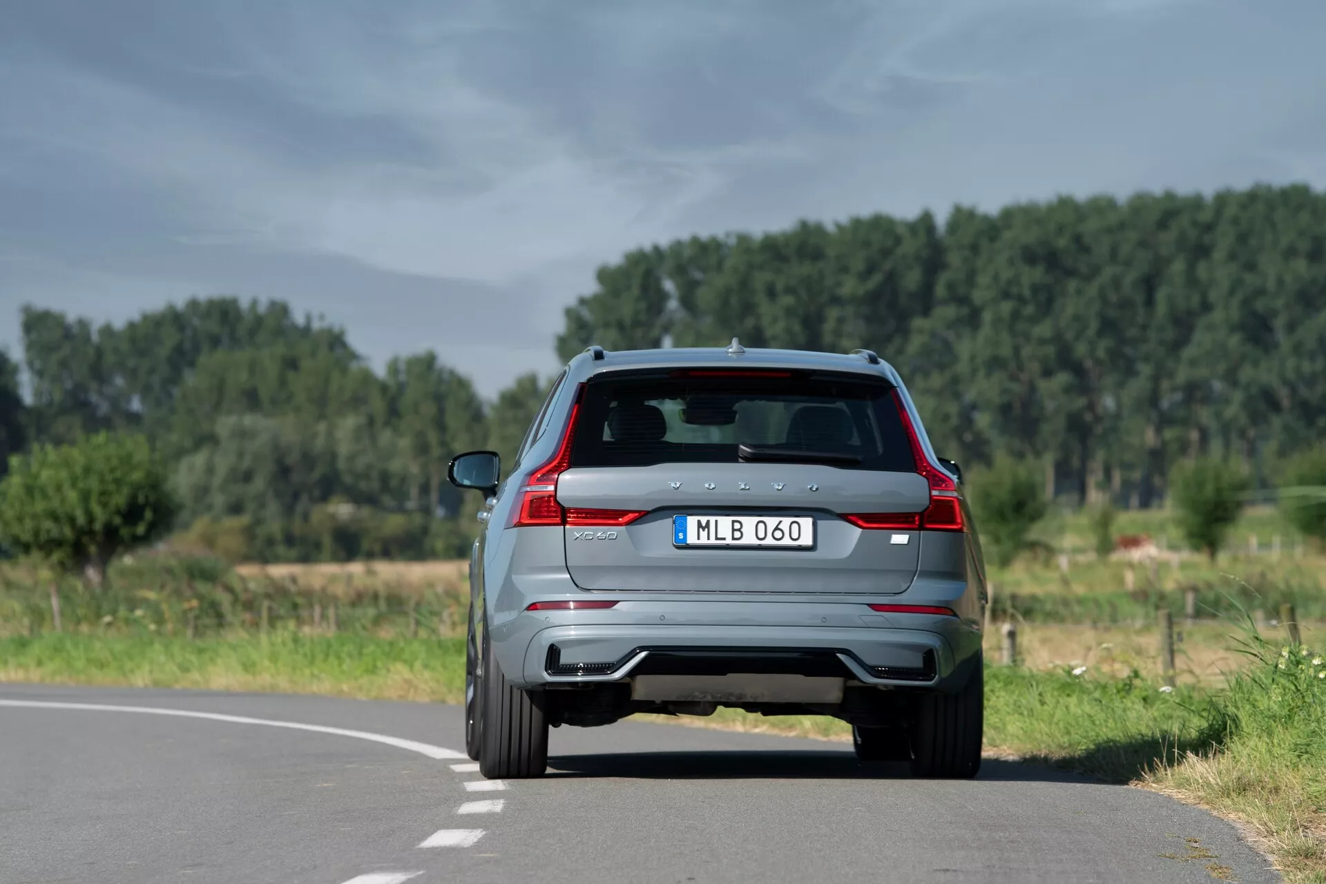Modern 2021-es Volvo XC60 SUV (2. generációs-facelift) hátsó nézete egy két sávos úton természetes, vidéki környezetben, jól látható hátsó lámpákkal, kettős kipufogóval, integrált féklámpás hátsó spoilerrel, hátsó kamerával, hátsó ablaktörlővel és parkolóérzékelőkkel. Kissé a középső sáv jelzése jobb oldalán helyezkedik el, tiszta égbolttal és fákkal a háttérben.