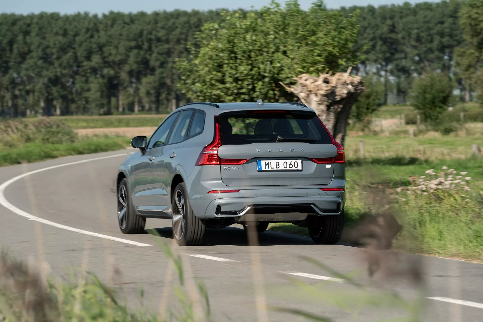 Háromnegyedes hátsó nézet egy 2021-es modern, metál szürke Volvo XC60 SUV-ról egy vidéki úton mozgás közben, ami jellegzetes hátsó lámpákat, kettős kipufogórendszert, hátsó ablaktörlőt, jellegzetes hátsó spoilert, integrált érzékelőket a hátsó lökhárítón, központilag elhelyezett rendszámtáblát, könnyűfém kerekeket, éles karaktervonalat az oldalon, fényvisszaverő ablakkereteket és tető sínrendszert mutat. Elmosódott növényzettel és tiszta égbolttal a háttérben a mozgást és a vidéki környezetet jelzi.