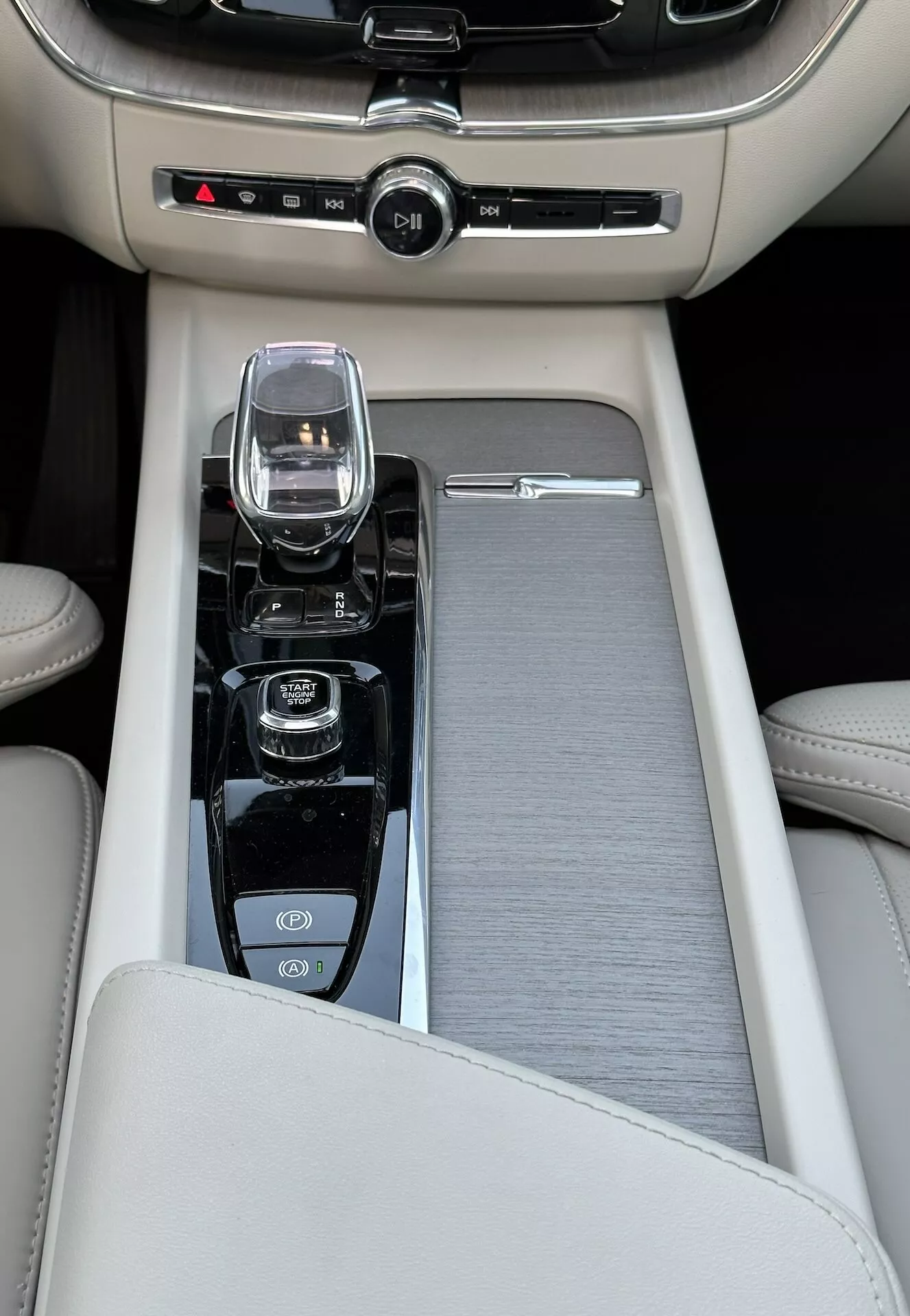 Közeli nézet a középkonzolra egy 2021-es Volvo XC60 Recharge plug-in hibridben, bézs bőrkarfával, csúszó kefélt fém díszítéssel, modern infotainment vezérlővel, kristályüveg sebességváltóval automatikus váltóhoz, és vezérlőkkel az indítás/leállítás motorhoz, tapadásgátló kikapcsolásához és a vezetési dinamika mód választásához, valamint kétzónás klímavezérléssel.