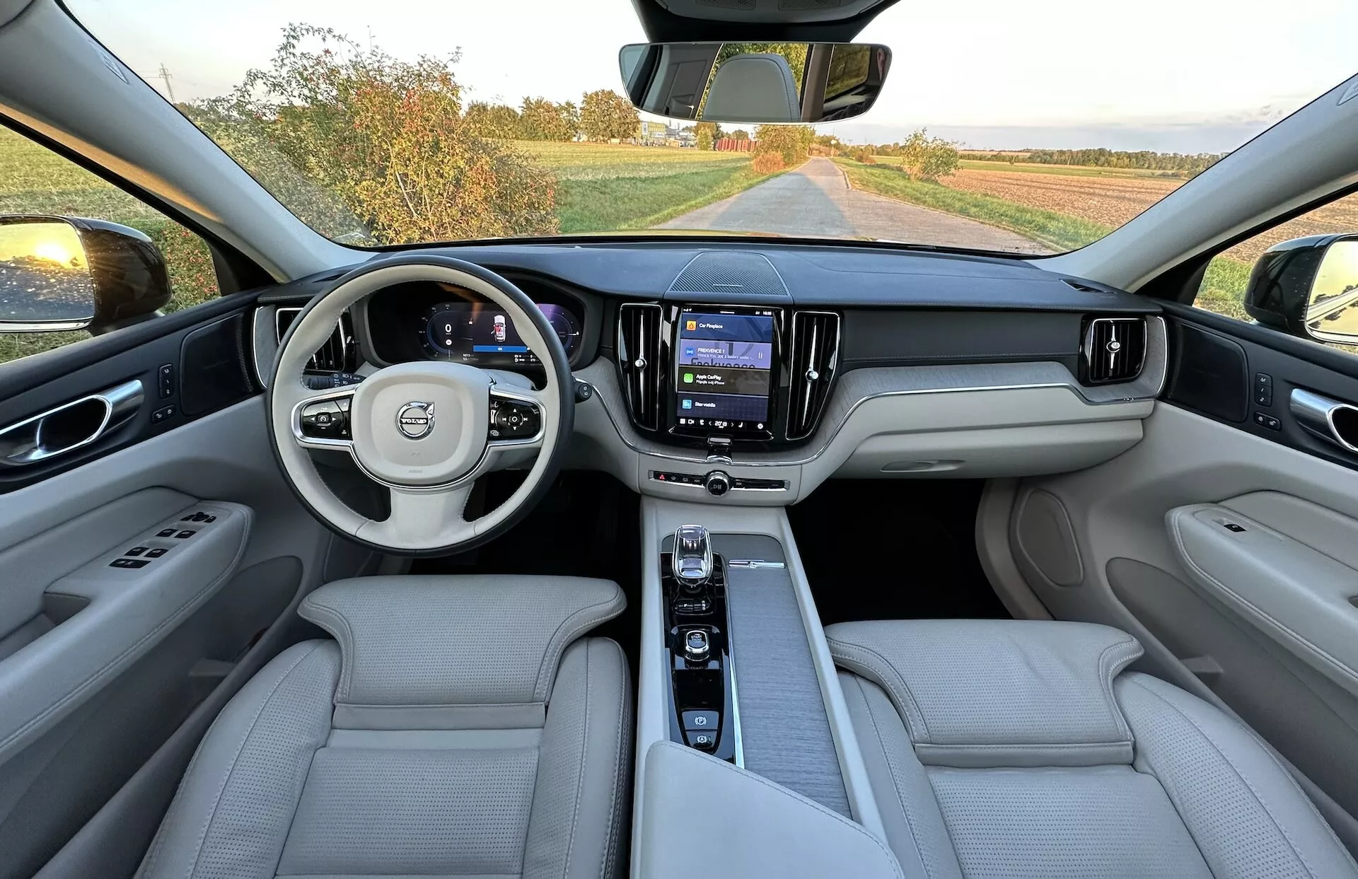 2021-es Volvo XC60 Recharge Plug-In Hybrid belső nézete a hátsó ülések területéről, az egész műszerfalat, multifunkcionális kormánykereket logóval, digitális műszerfalt, központi érintőképernyős infotainment rendszert és luxus világosszínű bőr üléseket mutatva.