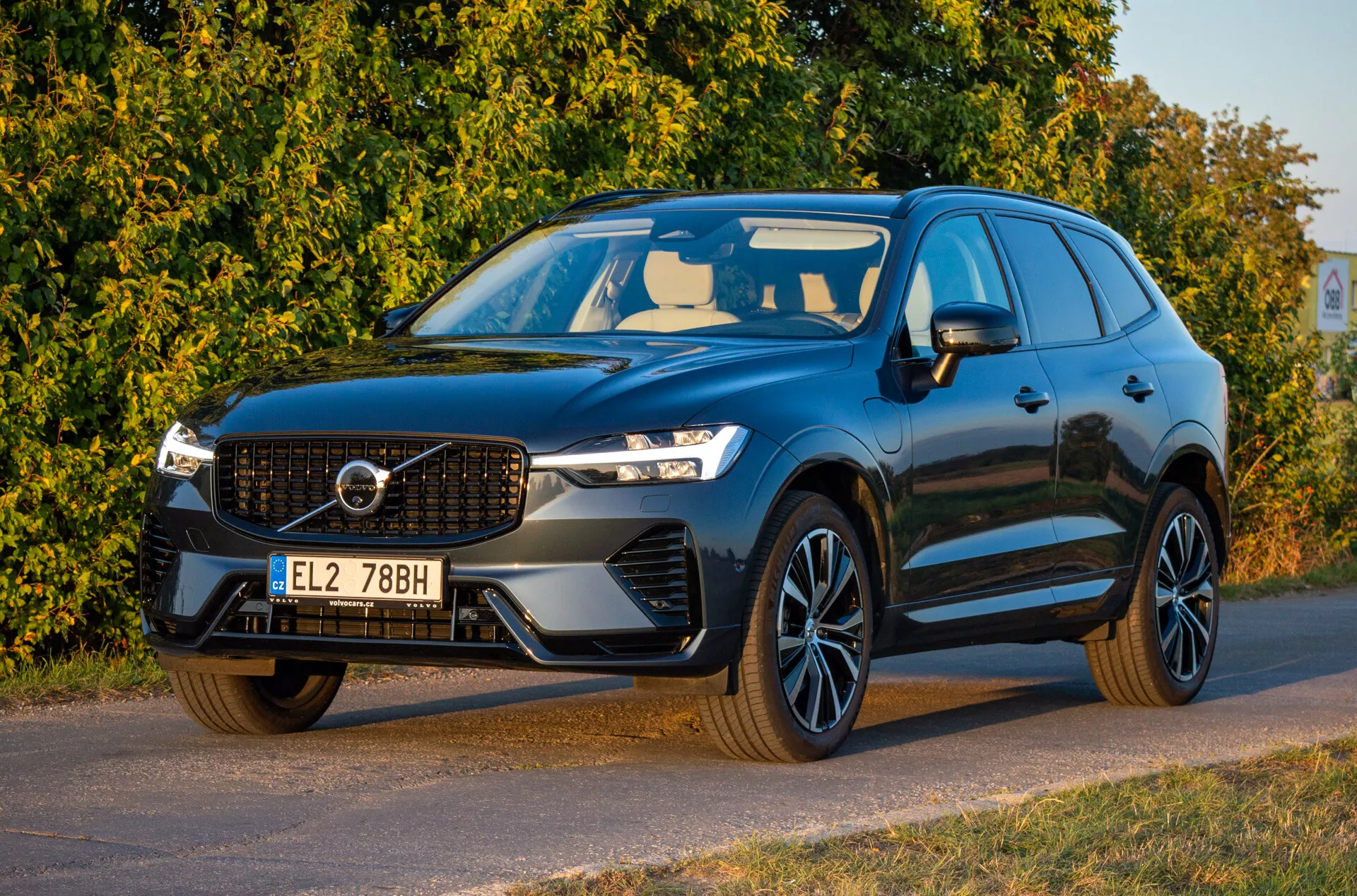 2021-es Volvo XC60 Recharge plug-in hibrid sötét színben, előlről háromnegyedes nézetben, amely bemutatja a LED fényszórókat, a jellegzetes hűtőrácsot, a sportos ötvözött kerekeket és az aerodinamikus tervezést, balra néző oldalirányban parkolva, az első rész és a sofőr oldala részletesen látható.