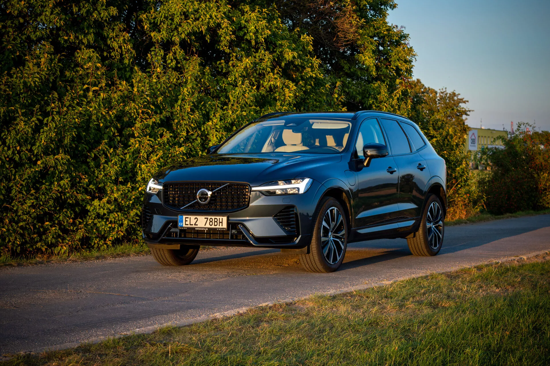 2021-es Volvo XC60 Recharge Plug-In Hybrid az út szélén parkolva késő délutáni meleg fényben, bemutatva az első hűtőrácsot, LED fényszórókat és sportos profilját a növényzet hátterével