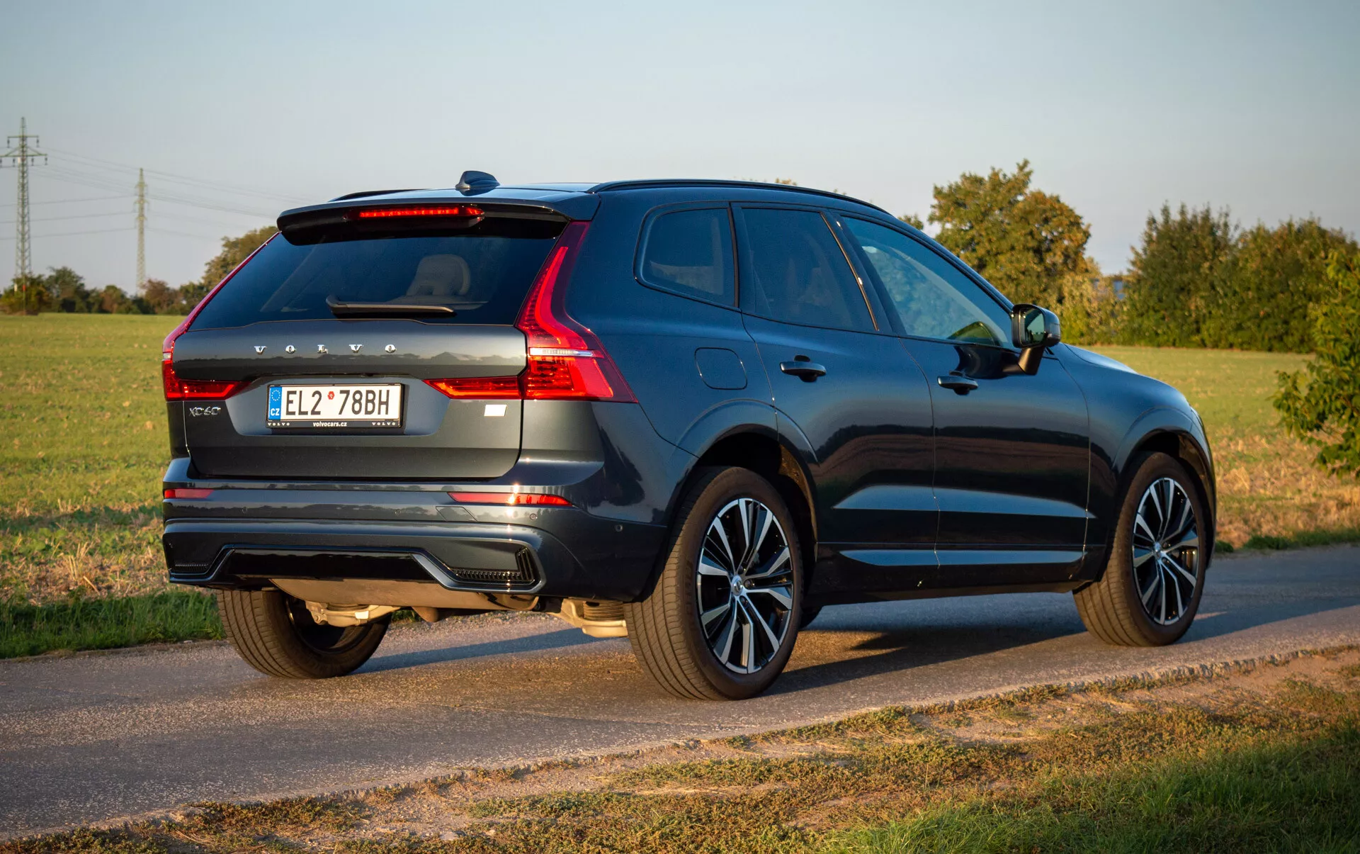 2021-es Volvo XC60 Recharge Plug-In Hybrid mélykék színben, aerodinamikus tervezéssel és modern jellemzőkkel, mint LED hátsó lámpák, ötvözött kerekek és kettős kipufogórendszer, vidéki úton parkolva tiszta ég alatt