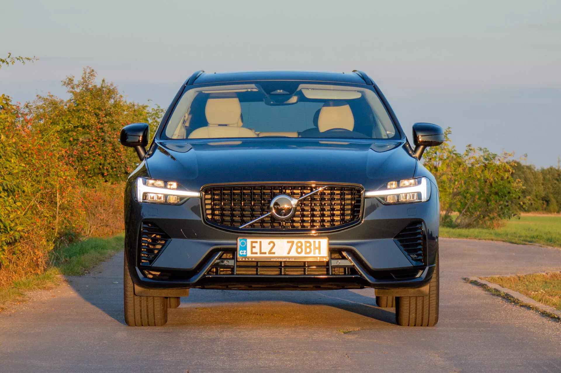 2021-es Volvo XC60 Recharge Plug-In Hybrid, amely modern tervezését mutatja be jellegzetes ráccsal, karcsú LED fényszórókkal és sportos első lökhárítóval vidéki háttér előtt késő délutáni meleg fényben