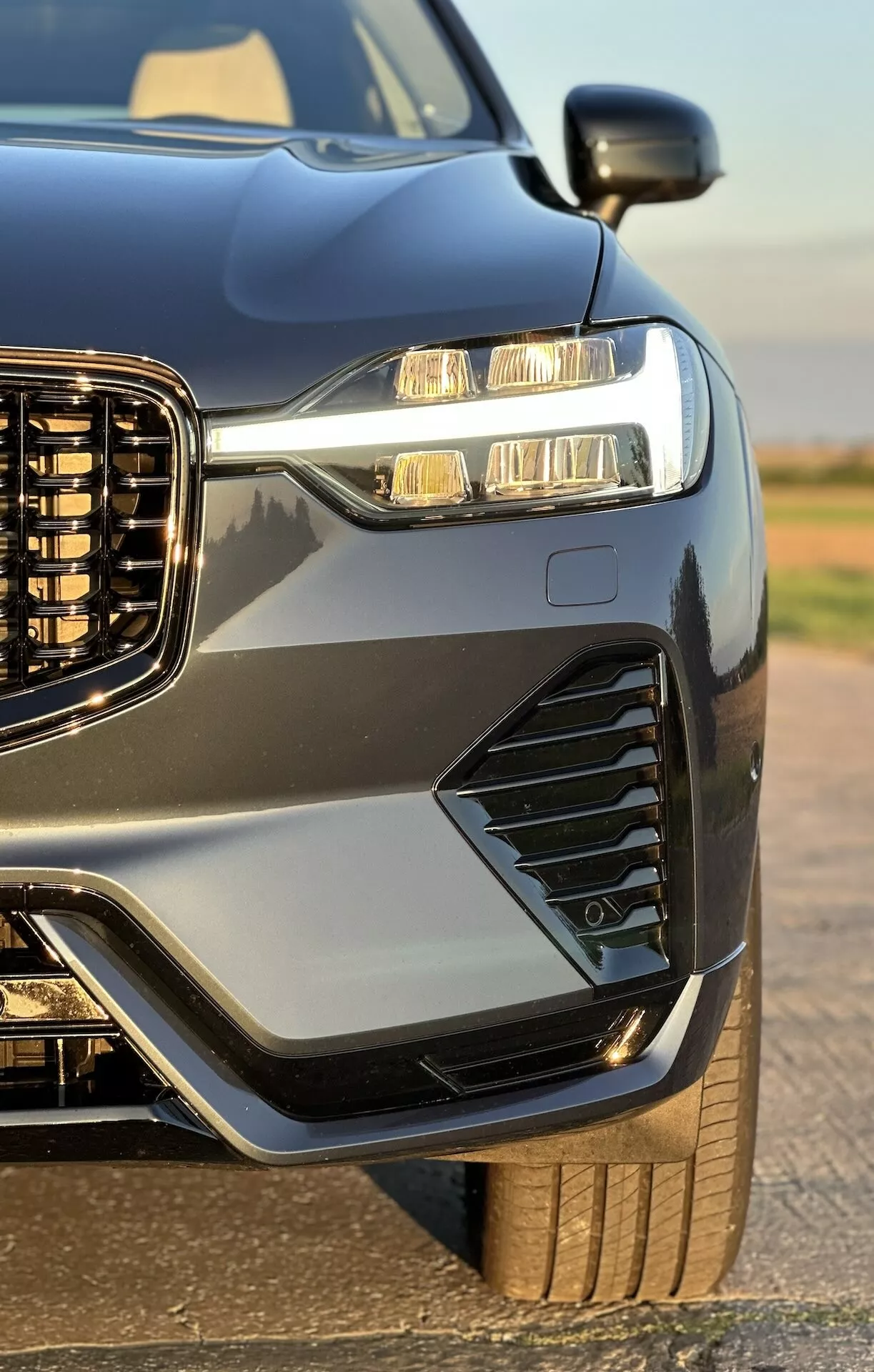 2021-es Volvo XC60 Recharge Plug-In Hybrid első negyedének közelképe, kiemelve a függőleges hűtőrácsot, LED fényszórókat nappali menetfényekkel, stílusos szellőzőnyílásokat és ködlámpákat, sportos motorháztetőt és sárvédőket, metál fényezést kontrasztos díszítőelemekkel, és a robosztus kerék valamint abroncs rövid betekintését, amit a jármű fényes bevonata emel ki.