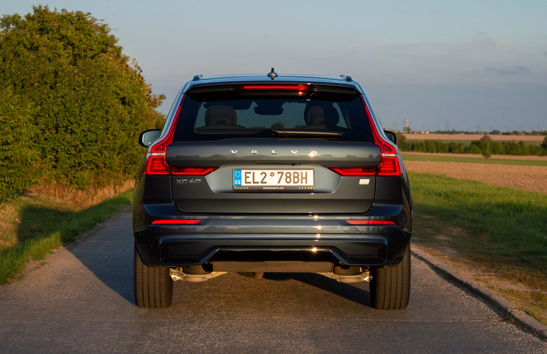 Kék 2021-es Volvo XC60 Recharge Plug-In Hybrid SUV hátsó nézete, függőleges hátsó lámpákkal, kettős kipufogóvégekkel, hátsó visszaverőkkel, hátsó ablaktörlővel, harmadik féklámpával, európai rendszámtáblával és tolatókamerával vidéki úton naplemente előtt