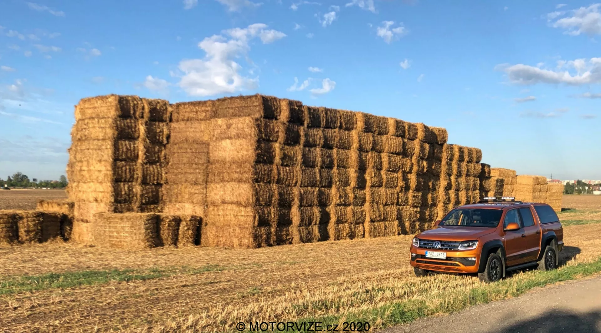 Egy 2016-os Volkswagen Amarok parkol nagy szalma bálák előtt vidéki környezetben. A háromnegyedes elülső perspektíva megmutatja az autó hűtőrácsát, fényszóróit, első lökhárítóját, motorháztetőjét, kerekeit, oldalsó tükröket és ajtókat. A kiemelkedő vízszintes rács és az éles, szögletes fényszórók növelik a jármű modern megjelenését. A sötétített ablakok, tetősínek és az ajtók körüli védőburkolatok sokoldalúságot és strapabírást sugallnak. A jármű szalma bálák háttere előtt helyezkedik el, utalva annak vidéki közlekedési vagy mezőgazdasági használatára. A reggeli vagy késő délutáni fény meleg árnyakat vet és kiemeli az autó színét és dizájn részleteit. 'MOTORVIZE.cz 2020' vízjellel.