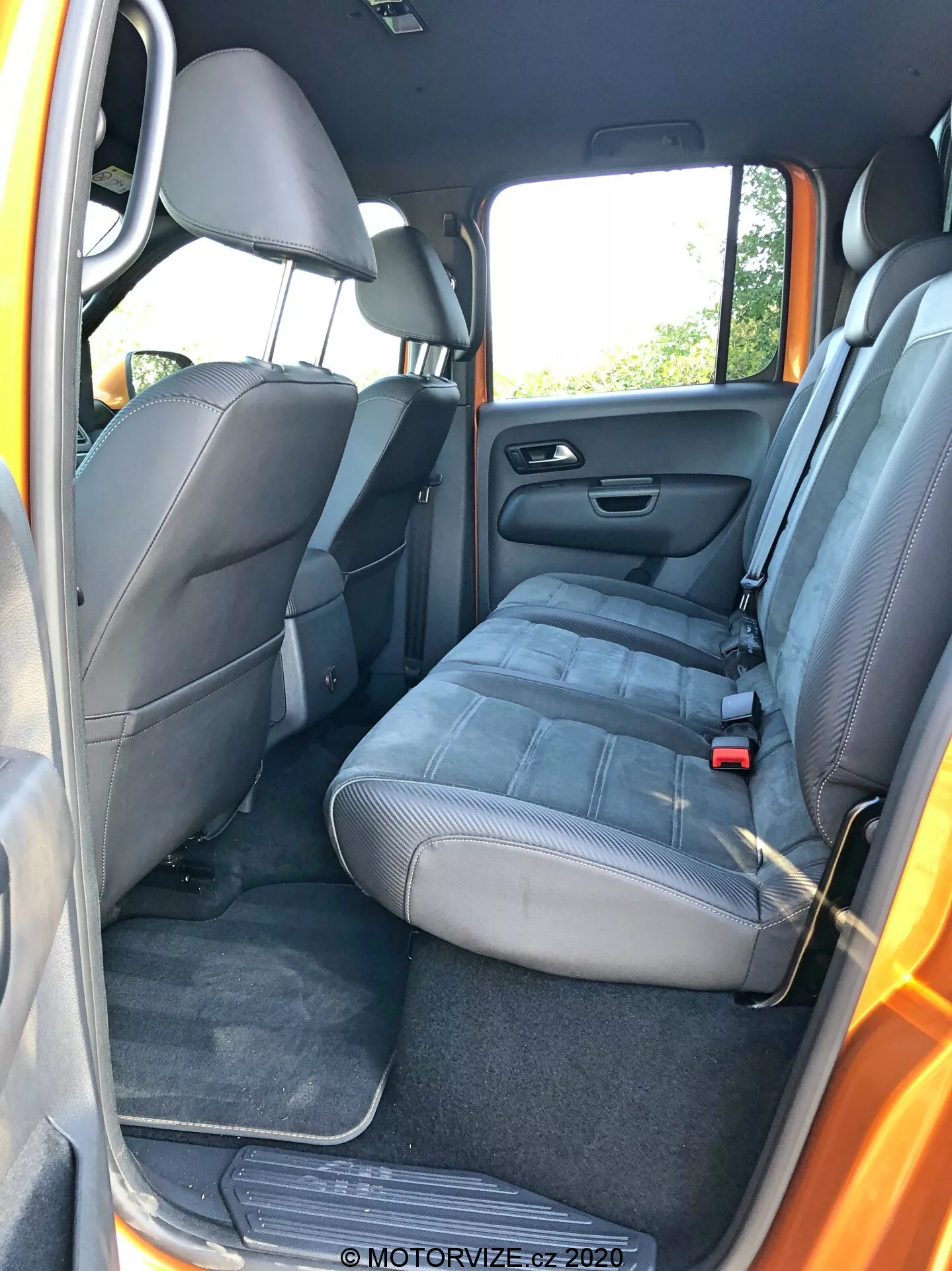 Belső nézet a VW-Nutzfahrzeuge Amarok 2016-ról a hátsó utas ajtajának szögéből; három utas számára alkalmas pad-stílusú hátsó ülés szövet kárpitozással, fejtámlákkal, biztonsági övekkel (középen csípőöv, oldalt hárompontos övek), nyitott ajtó, amely hangszórót, tárolót és elektromos ablakvezérlőt mutat; szőnyegpadló és lehajtható hátsó ülések; tárolózseb az első utasülés hátulján; standard autós funkciók kiemelkedő csúcstechnikai elemek nélkül; természetes nappali fény.