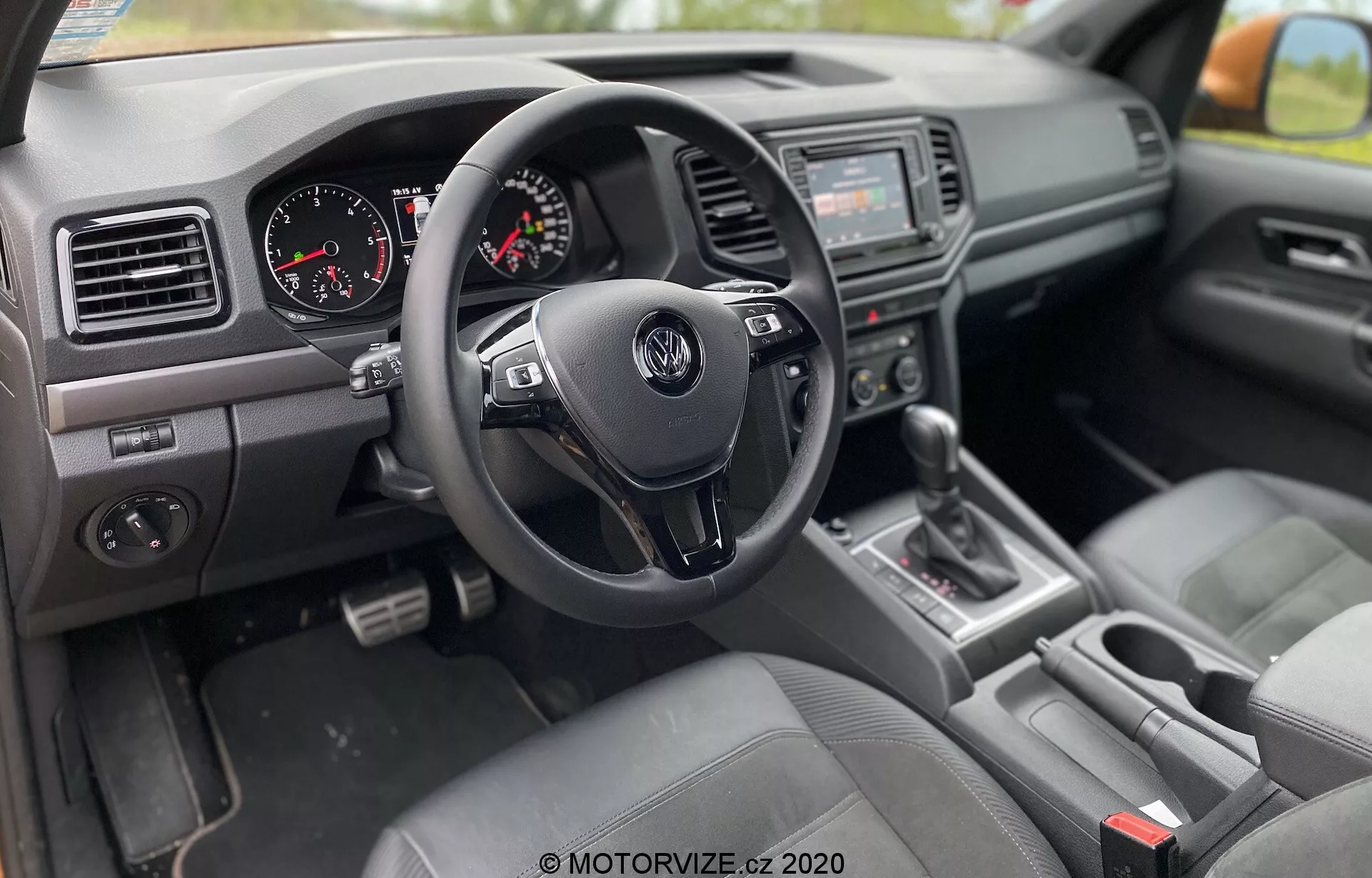 A 2016-os VW Nutzfahrzeuge Amarok belső nézete az utas oldaláról, jobbkormányos kialakítással, háromágú Volkswagen kormánykerékkel, kezelőgombokkal, műszerfallal analóg sebesség- és fordulatszámmérővel, középkonzollal audiorendszerrel és infotainment képernyővel, 'P' helyzetben lévő sebességváltóval, szövettel bevont ülésekkel, szellőzőnyílásokkal és texturált műanyagot tartalmazó műszerfalal, valamint a vezető ajtajának paneljével elektromos ablakok és tükörállítás vezérlési lehetőségeivel.