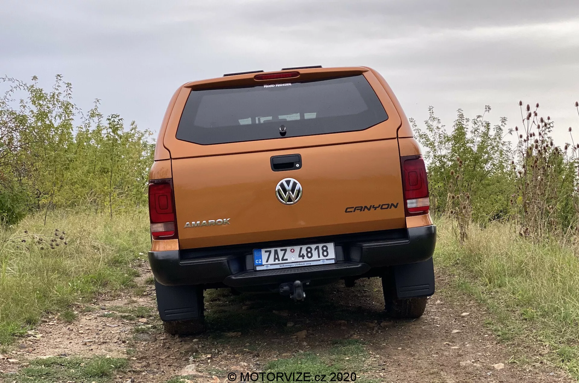 Professzionális hátsó nézet egy 2016-os Volkswagen Amarok pick-up teherautóról alacsony szögből, kiemelve a robusztus hátfalat 'AMAROK' márkajelzéssel, középen Volkswagen logóval, 'CANYON' felszereltségi szint jelzővel, rendszámtábla, részletes hátsó lámpák, lökhárító parkolószenzorokkal és lépcsővel, vonórúd, egy burkolatlan úton parkolva, ami terepjárási képességet jelez.