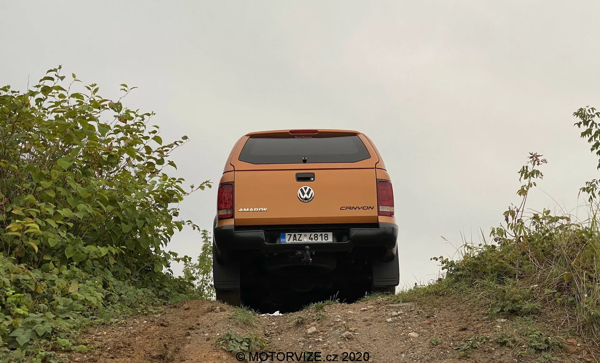 Egy narancssárga Volkswagen crossover vagy SUV hátsó nézete enyhén alacsony szögből, amely egy kis domb tetején készült. A jármű hátsó design elemei, például a csomagtér ajtó, a hátsó ablak, a hátsó lámpák, a márka és a modell jelzése, amelyen a 'Volkswagen' és a 'CROSSOVER' felirat olvasható, valamint a hátsó lökhárító jól láthatóak. A rendszámtábla látható, de konkrét részletek nem közöltek. A csomagtér ajtó kiemelkedő hajtással és vonalvezetéssel rendelkezik, a rendszámtábla alatt esetleg egy fogantyú vagy mélyedés található. A hátsó lámpák oldalra fordulnak, és tartalmazzák az irányjelzőket, a tolatólámpákat és a féklámpákat. A hátsó lökhárító alsó fekete része védő díszlécet jelez a terepi használathoz. A jármű növényzet szegélyezi, ami vidéki vagy terepi környezetre utal. A talaj természetes, ami összhangban van a jármű terepjáró képességeivel. A jobb alsó sarokban egy 'MOTORVIZE.cz ©2020' feliratú vízjel található.