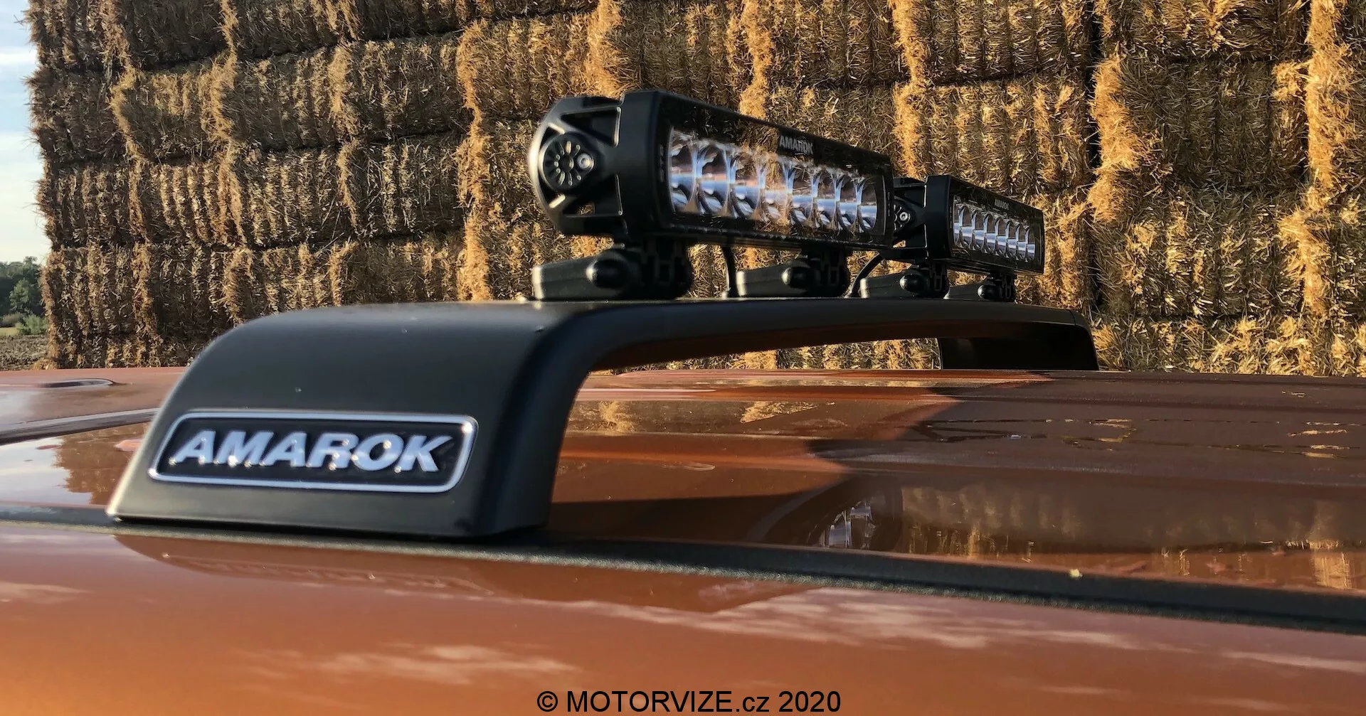 Közelkép egy 2016-os Volkswagen Amarok pickupról, amelyen egy utólagos LED világítósáv van a tetőn elhelyezve. A világítósáv fekete burkolatában láthatóak az egyes LED elemek, a 'AMAROK' felirat pedig egy szellőző burkolaton található. A kinti felvételen látható a világítósáv tükröződő lencséje és szalmabálák a háttérben, jelezve a vidéki környezetet.
