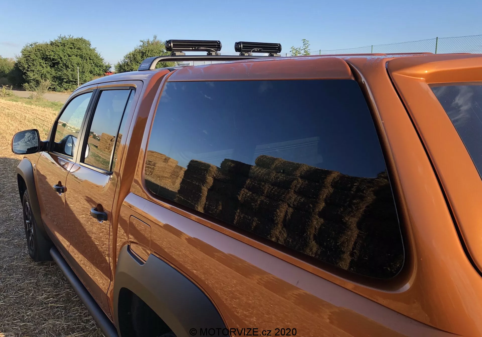 Közeli, oldalnézeti felvétel egy narancssárga pickupról, amely főként a fülke felső hátsó részét és a plató egy részét mutatja. A jellemzők közé tartozik a tiszta oldalsó profil, a hátsó utasablakra összpontosítva, amely egy vidéki környezetet tükröz szénabálákkal, tetőcsomagtartó extra rakomány számára, modern, enyhén sötétített ablaktervezéssel, fényes fényezéssel, jellegzetes narancssárga színnel, látható jelvénnyel, részleges oldalsó tükörrel és meleg napfénnyel, amely a kora reggeli vagy késő délutáni időpontot sugallja.