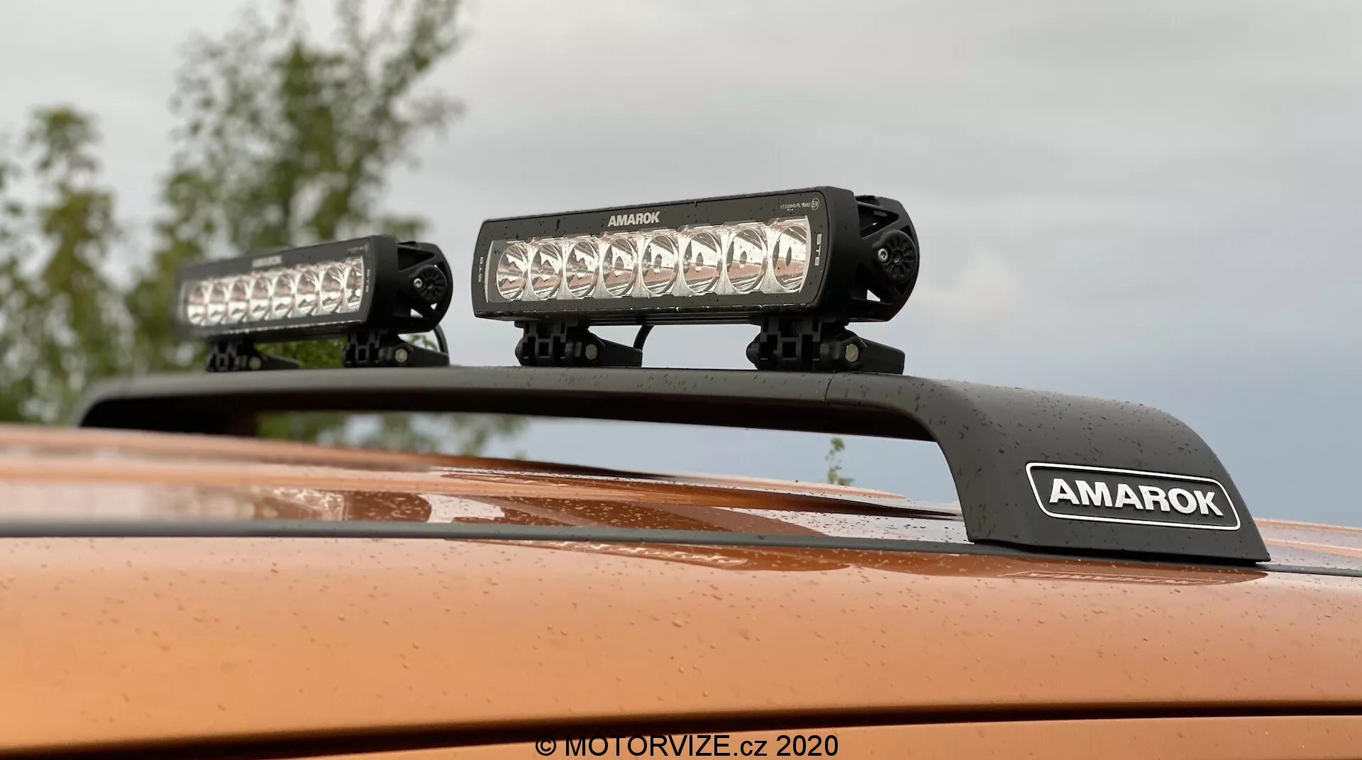 Felfelé fókuszált közeli kép egy 2016-os Volkswagen Amarok tetejéről, amely utólagosan felszerelt tetővilágítást, két nagy intenzitású LED fényrudat mutat az 'AMAROK' márkájú tartón, fémes narancs festék kivitelben; ideális terepjáró járműtulajdonságokhoz vagy járműkiegészítőkhöz.