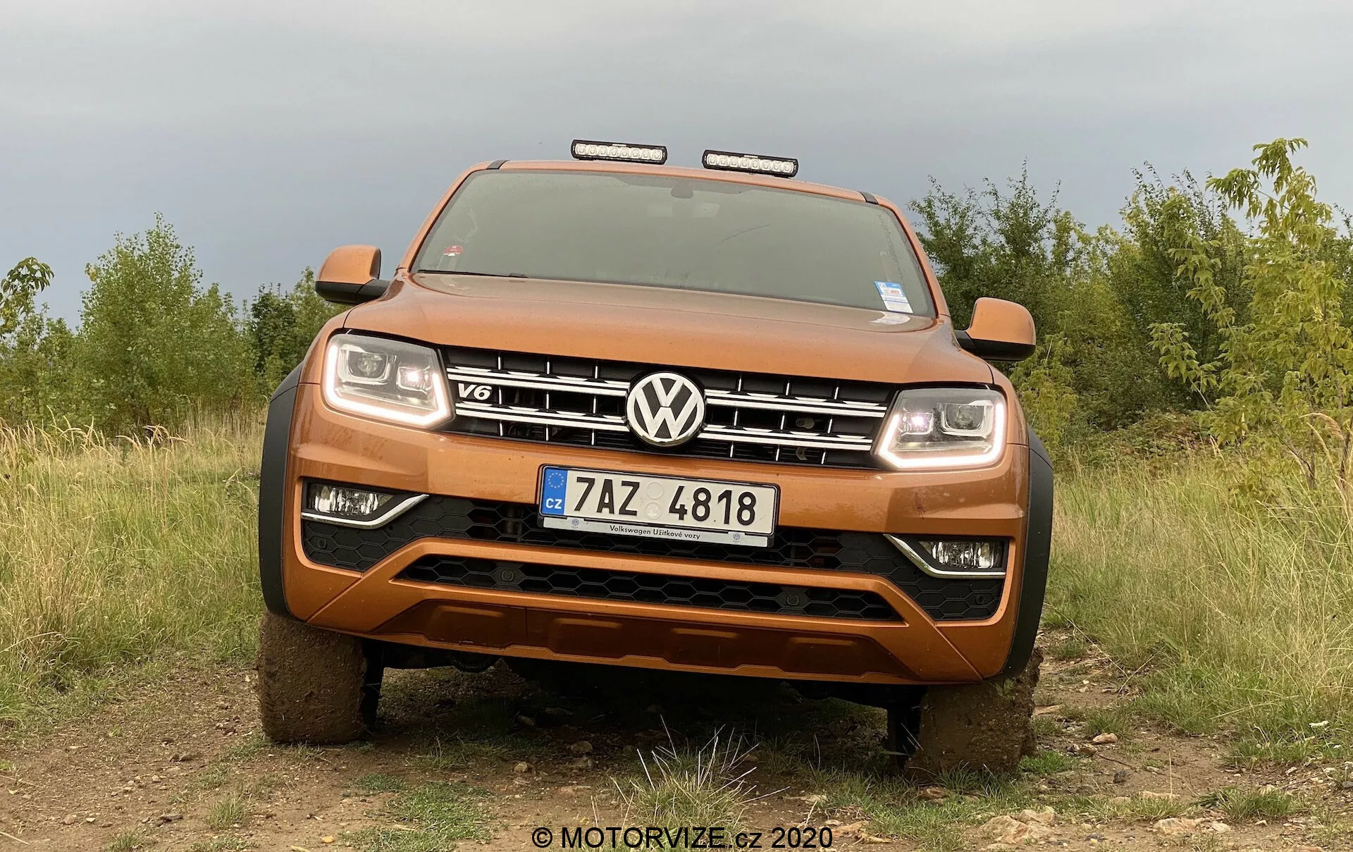 Fémdobozos narancssárga Volkswagen Amarok pickup előnézete, amely robusztus tartását és terepjáró képességeit mutatja, feltűnő hűtőrácsával a VW emblémával és a V6 felirattal. A kép alacsony szögből készült, és a jármű teljes elülső szélességét mutatja, beleértve a fényszórókat és a kiegészítő terepjáró lámpákat, természetes háttérrel, mezővel és fákkal, amelyek kiemelik a pickup robusztusságát. A látható jellemzők közé tartozik a masszív első lökhárító, a csúszólemez, az aktivált fejlett világítási rendszer és a cseh rendszám.