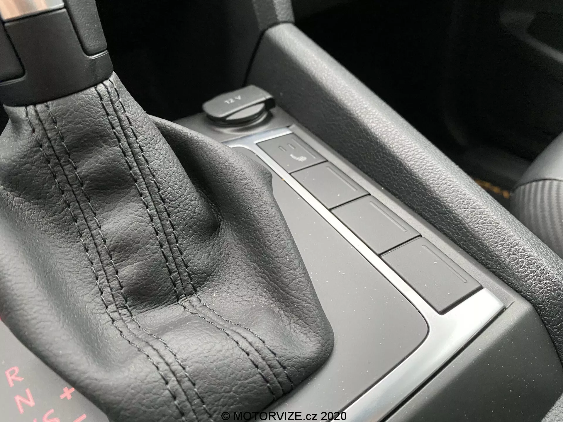 Közelkép egy VW Nutzfahrzeuge Amarok 2016 középkonzoljáról, a sebességváltó területére fókuszálva. A középpontban a sebességváltó kar áll, amely fekete bőrrel van bevonva, látható varratokkal. A 'PRNDS' betűk a váltó mögött automata sebességváltóra utalnak. Jobbra négy négyzet alakú gomb található, amelyek valószínűleg különböző autófunkciókat irányítanak. A gombok mögötti terület egy kis tárolóhely lehet. Az anyagok és a kivitelezés minősége magas szintű kézművesmunkát sejtet. A bal felső sarokból érkező környezeti fény kiemeli a részleteket.