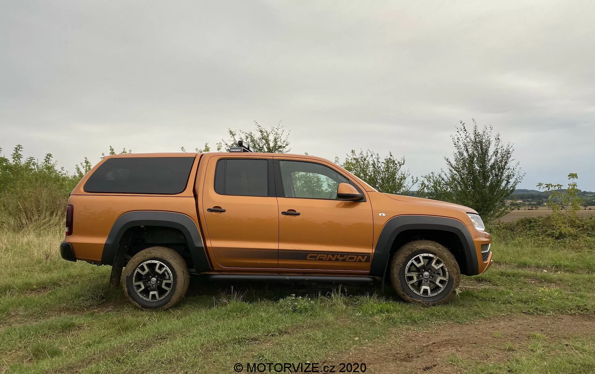 Narancssárga VW Nutzfahrzeuge Amarok 2016-os pickup autó oldalnézete, kemény héjú tónna fedéllel, egyenetlen füves terepen parkolva. A jármű magas földszegélyt, masszív kerekeket és gumiabroncsokat, valamint sokágú, ötvözetből készült dizájnt mutat, amely alkalmas terephasználatra. Látható jellemzők közé tartoznak az oldalsó tükrök, ajtókilincsek, kerékívek, fellépő, tágas négyajtós kabin, az első keréknél található jelvény és a hátsó kerék felett lévő díszszint megjelölése. A természetes fény és az egyenletes szög kiemeli a jármű részleteit anélkül, hogy erős árnyékokat vetne, hangsúlyozva annak terepjárási képességeit és biztos rakodási tárolóját.