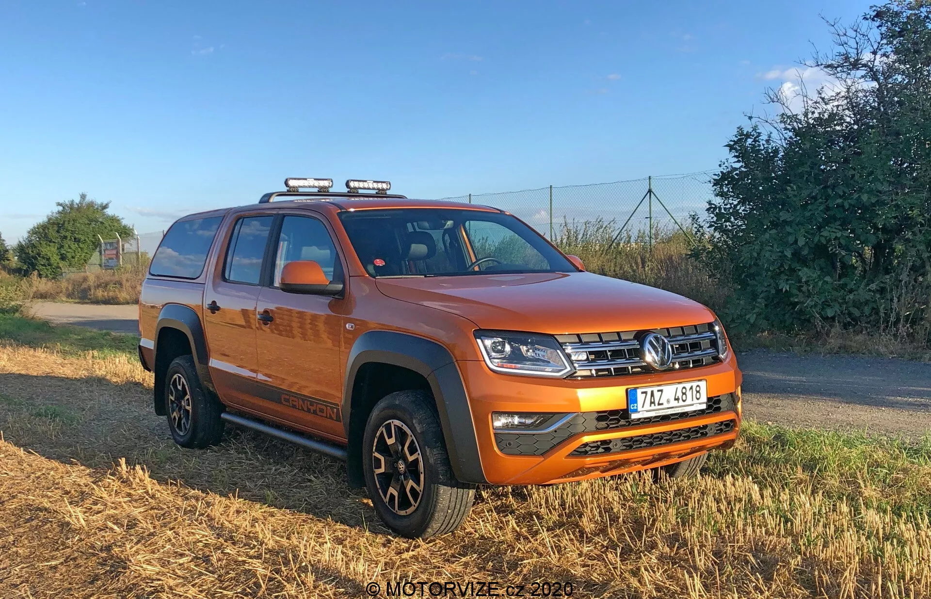 Kép egy vibráló narancssárga, négyajtós VW Nutzfahrzeuge Amarok 2016 pickup teherautóról, amely háromnegyedes szögből van fényképezve, a jármű elejét és oldalát mutatva. A kép kiemeli a vízszintesen bordázott elülső rácsot, a komplex LED fényszórókat, a tetőre szerelt segédfényeket, a szilárd lökhárítót ködlámpákkal, az alvázvédőt, a motorháztető emelt középső részét, a kerékíveken lévő fekete védőborítást, a többcélú gumikkal felszerelt fekete kerekeket, az ajtók alatti fellépőt, az integrált irányjelzős oldalsó tükröket, az takaróponyvával fedett platót és a további tárolókapacitást biztosító tetősíneket.