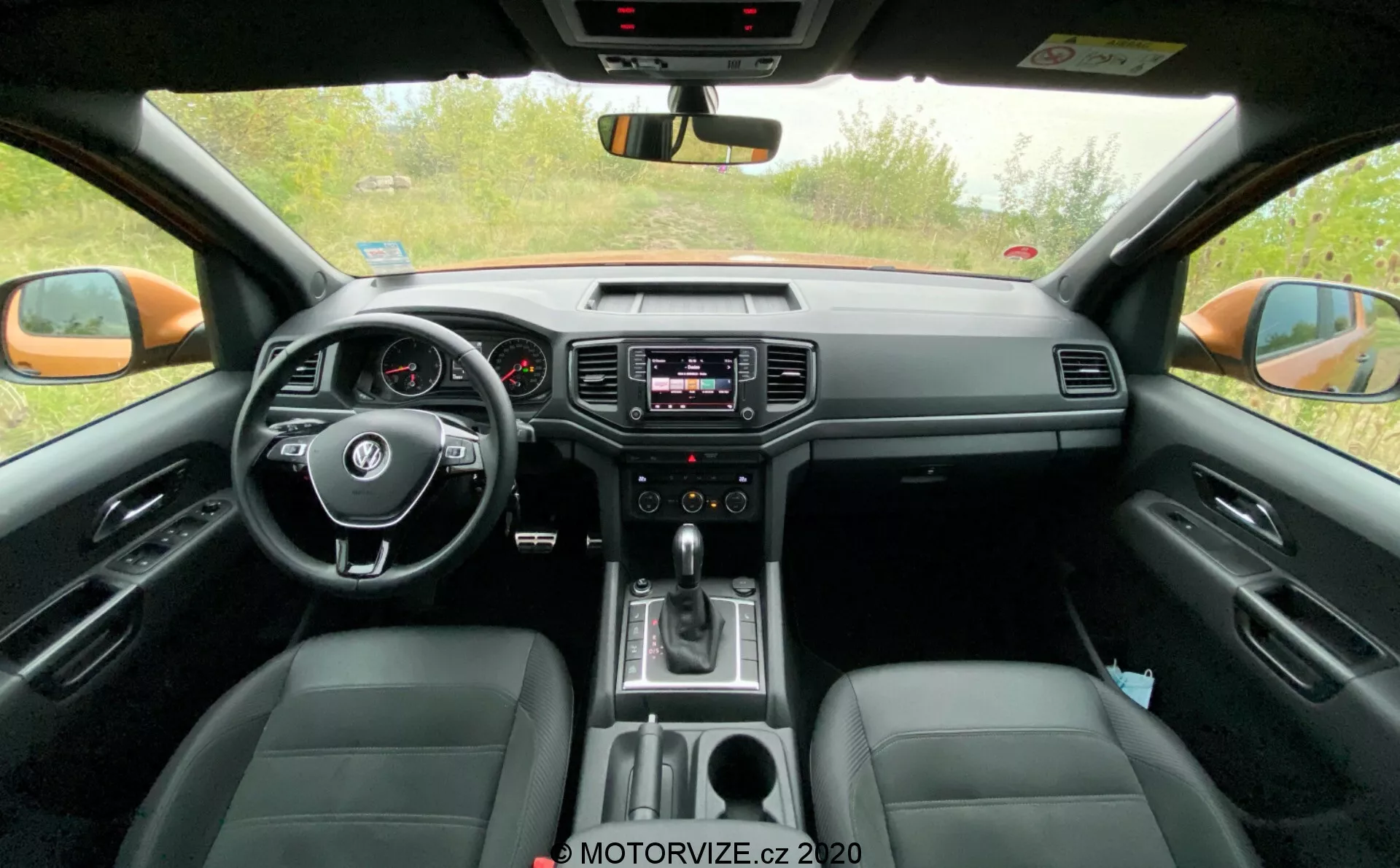 A 2016-os VW-Nutzfahrzeuge Amarok első belső kabinfotója. A hátsó ülés nézőpontjából készült kép középen található kormánykerékkel, rajta beépített vezérlőkkel, analóg műszercsoporttal, kis digitális kijelzővel, a műszerfal közepén elhelyezkedő infotainment rendszerrel, amely audio- és klímavezérlést kínál, valamint automata váltót jelző váltókarral és bőrülésekkel. A kép továbbá megjeleníti a visszapillantó tükröt, a külső visszapillantó tükröket és az első oldalablakokat, kiemelve a technológiai és kényelmi jellemzőket.